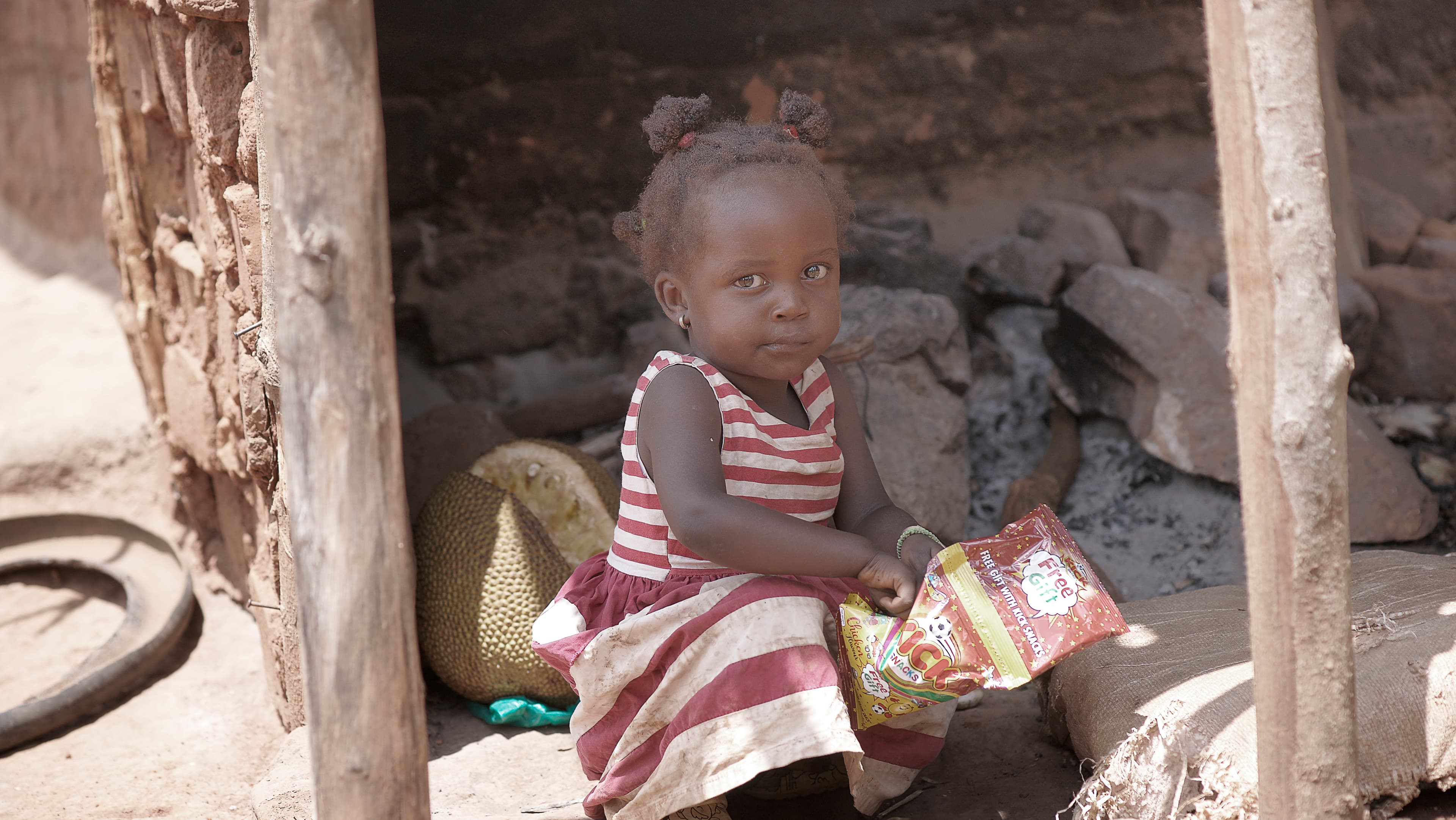 Toddler sitting outdoors near a wooden post, holding a food packet