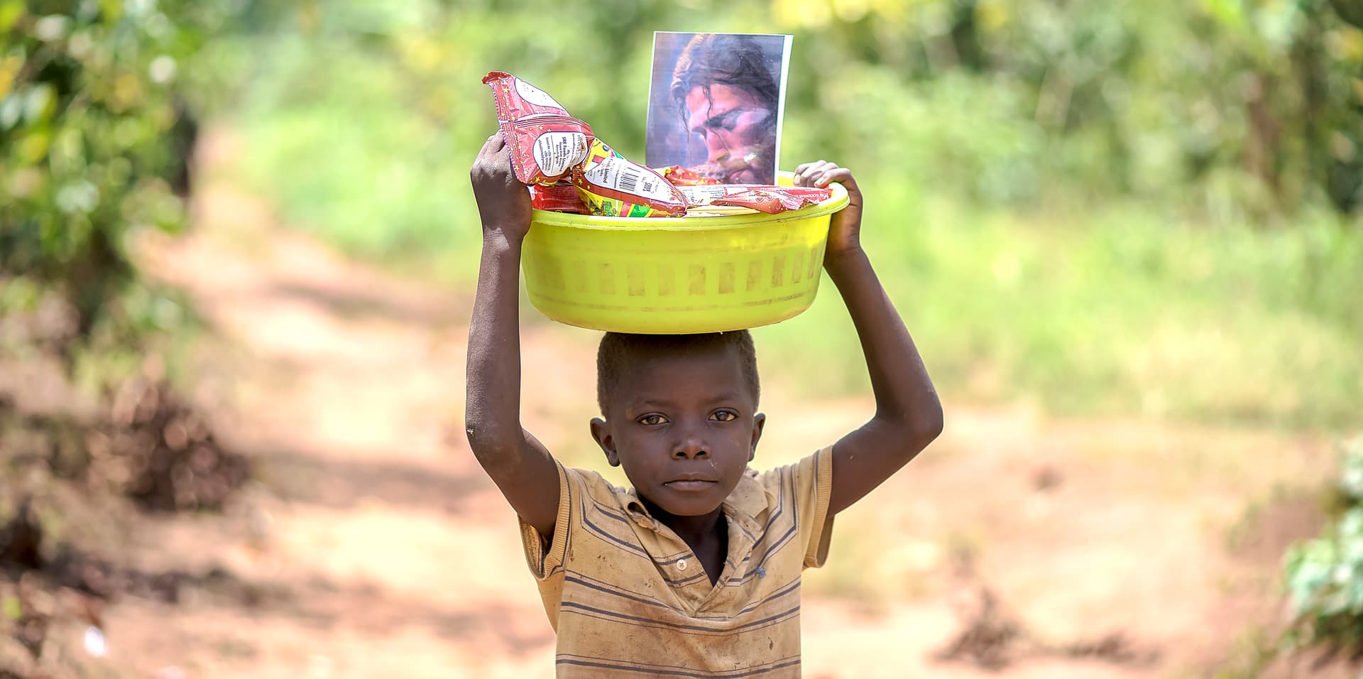 Child in a yellow shirt carrying a bright basin of food and a photo card on their head along a dirt path