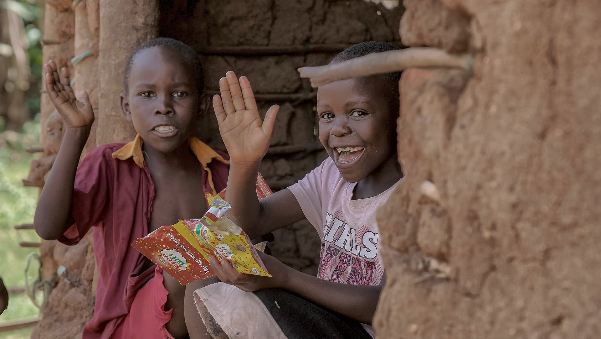 Two children sitting against a mud wall, one waving while holding a food packet