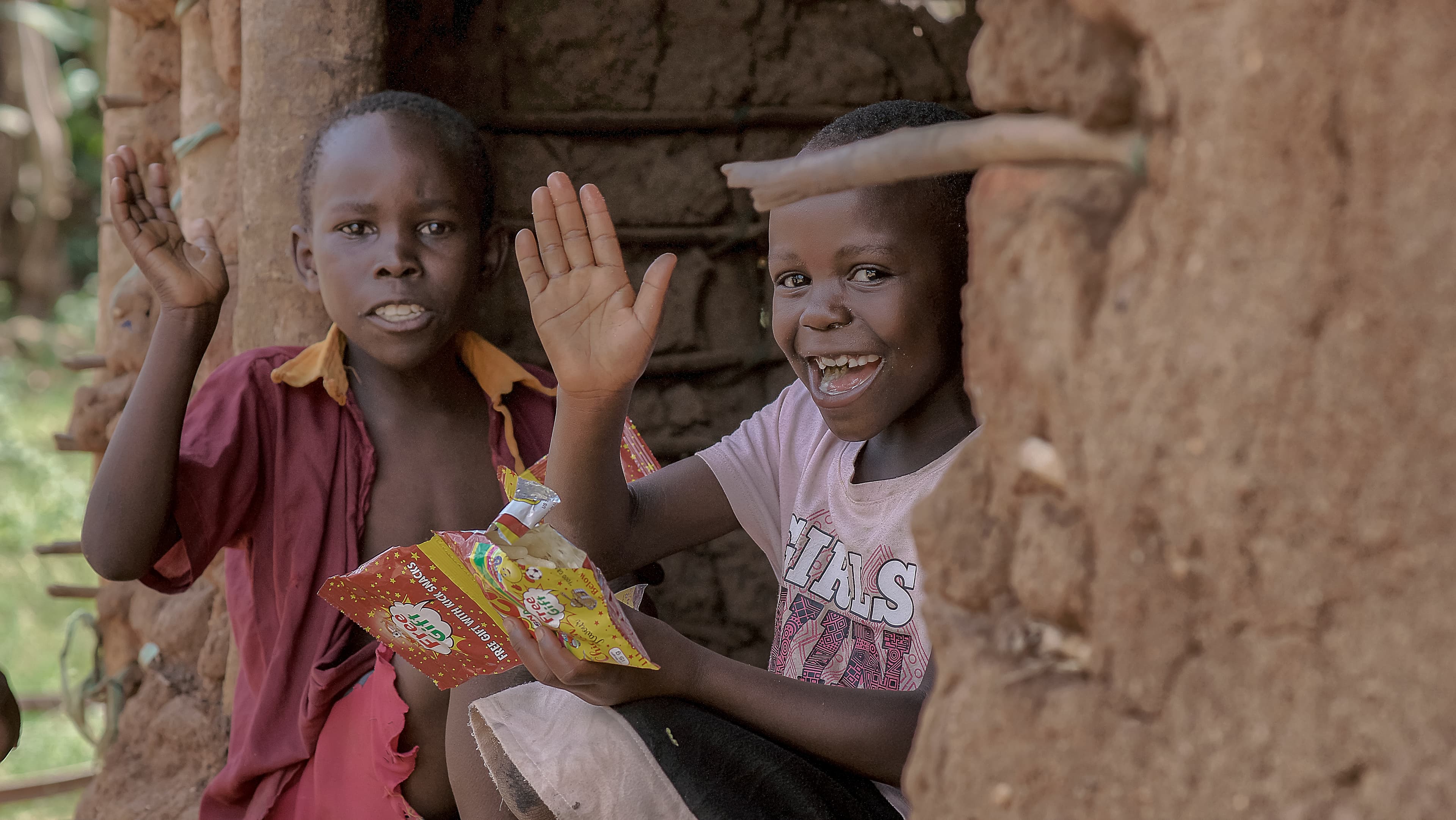 Two children sitting against a mud wall, one waving while holding a food packet
