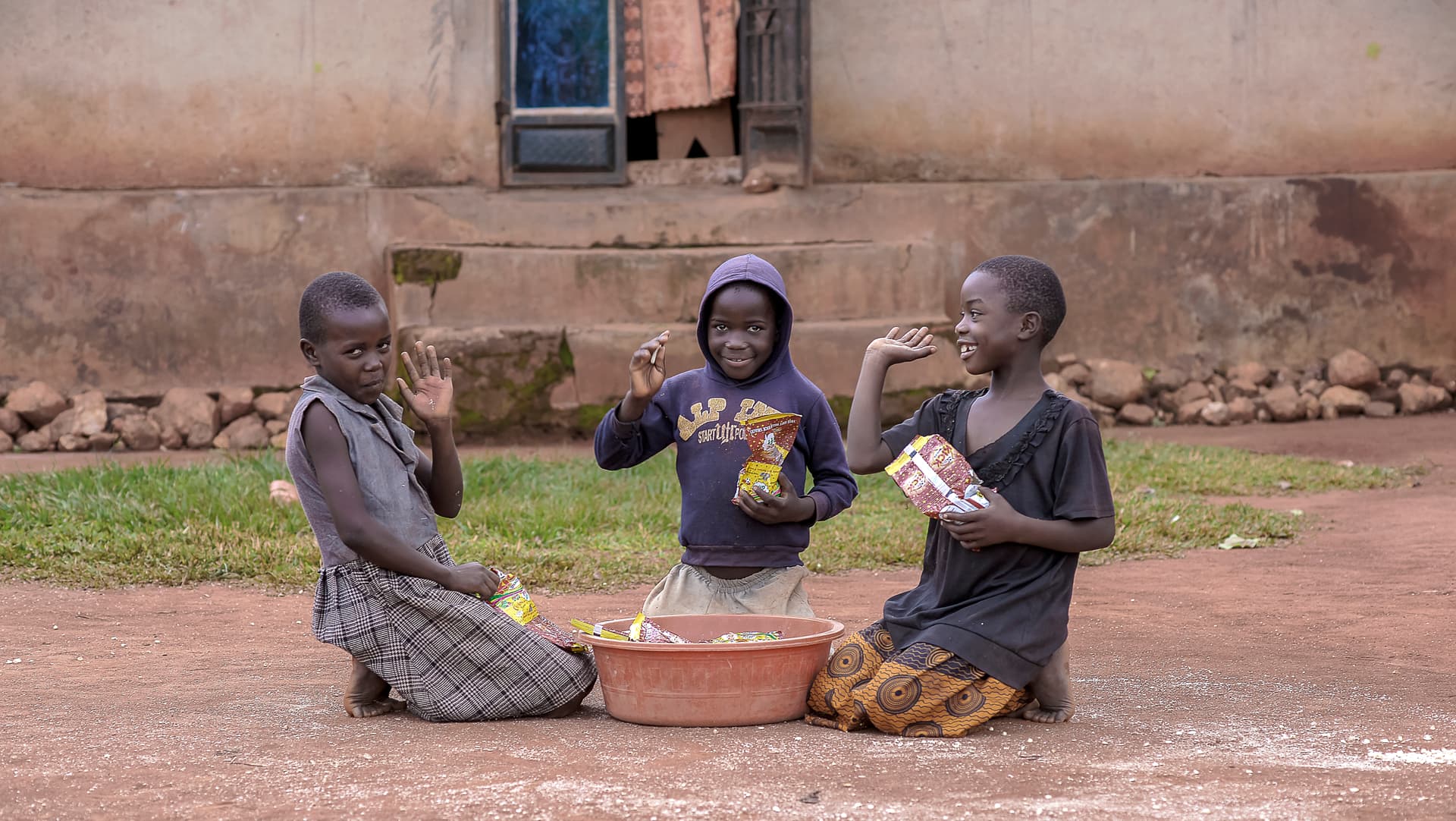Two children sitting together outdoors eating from a shared bowl on the ground