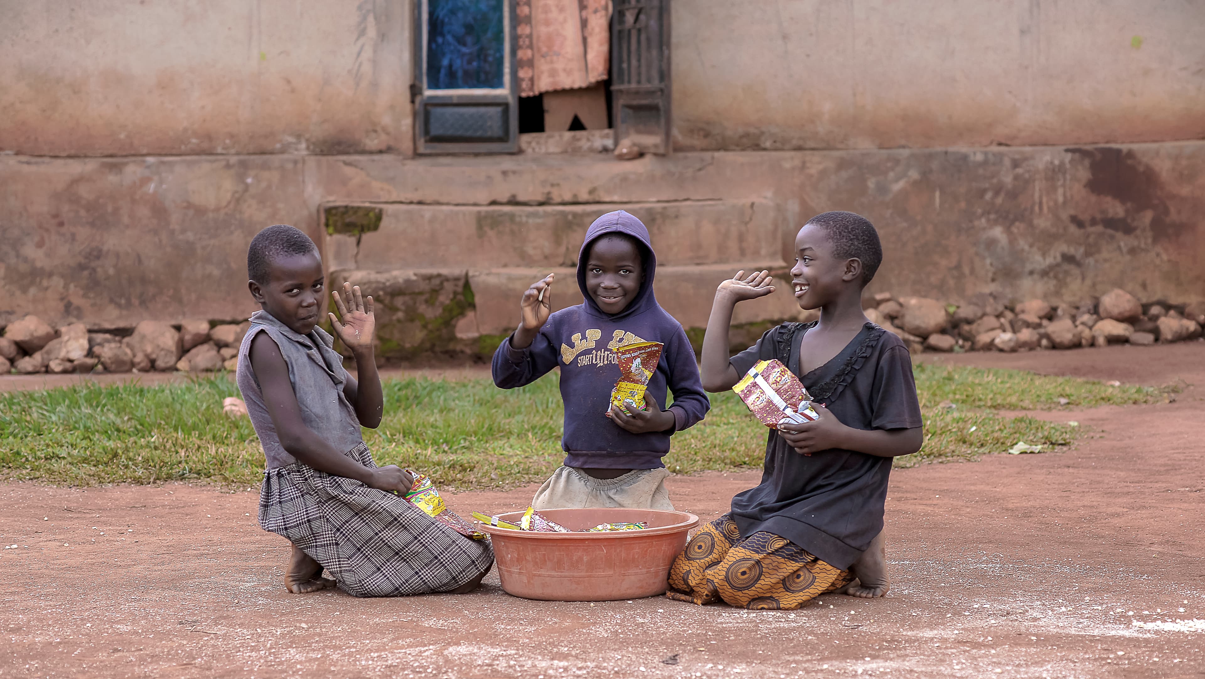 Two children sitting together outdoors eating from a shared bowl on the ground