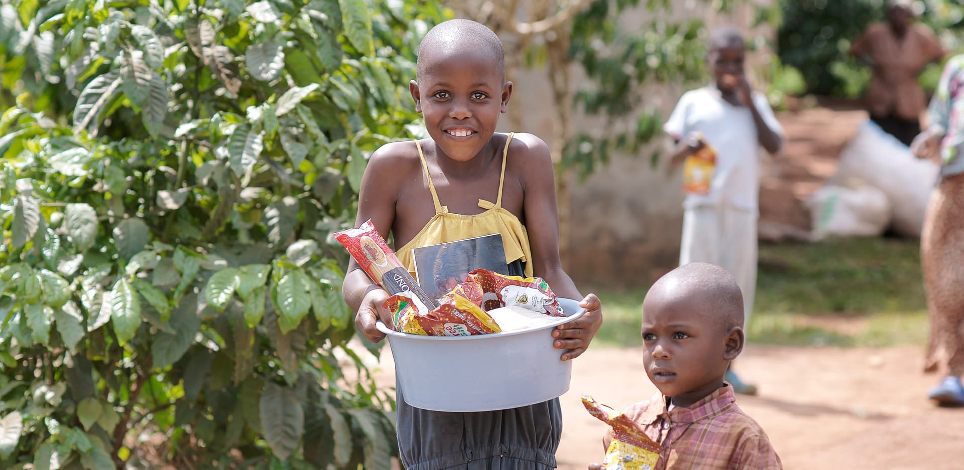 Girl and toddler outdoors, the girl holding a white basin of food supplies