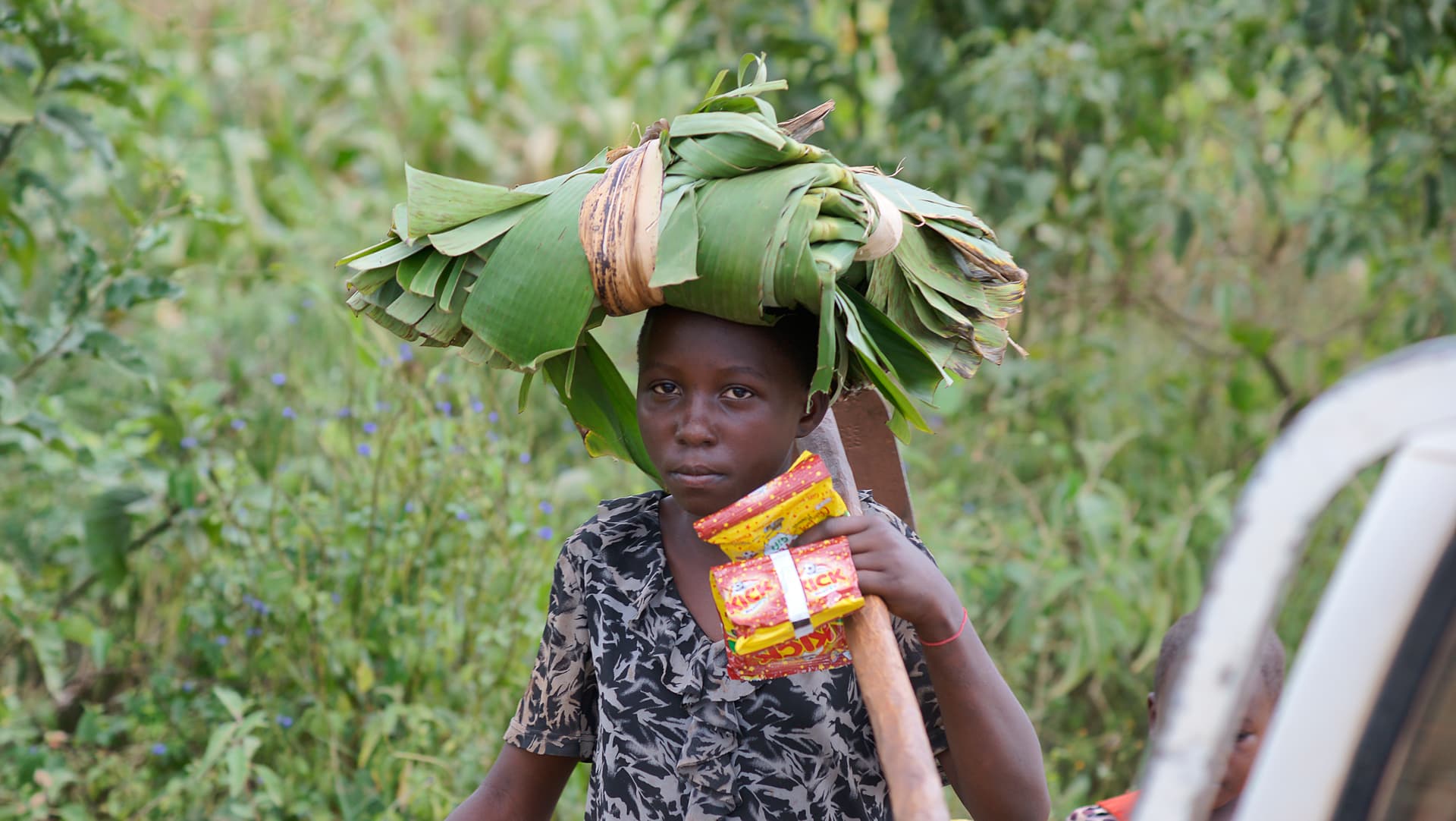 Child carrying a large bundle of banana leaves and food on their head outdoors