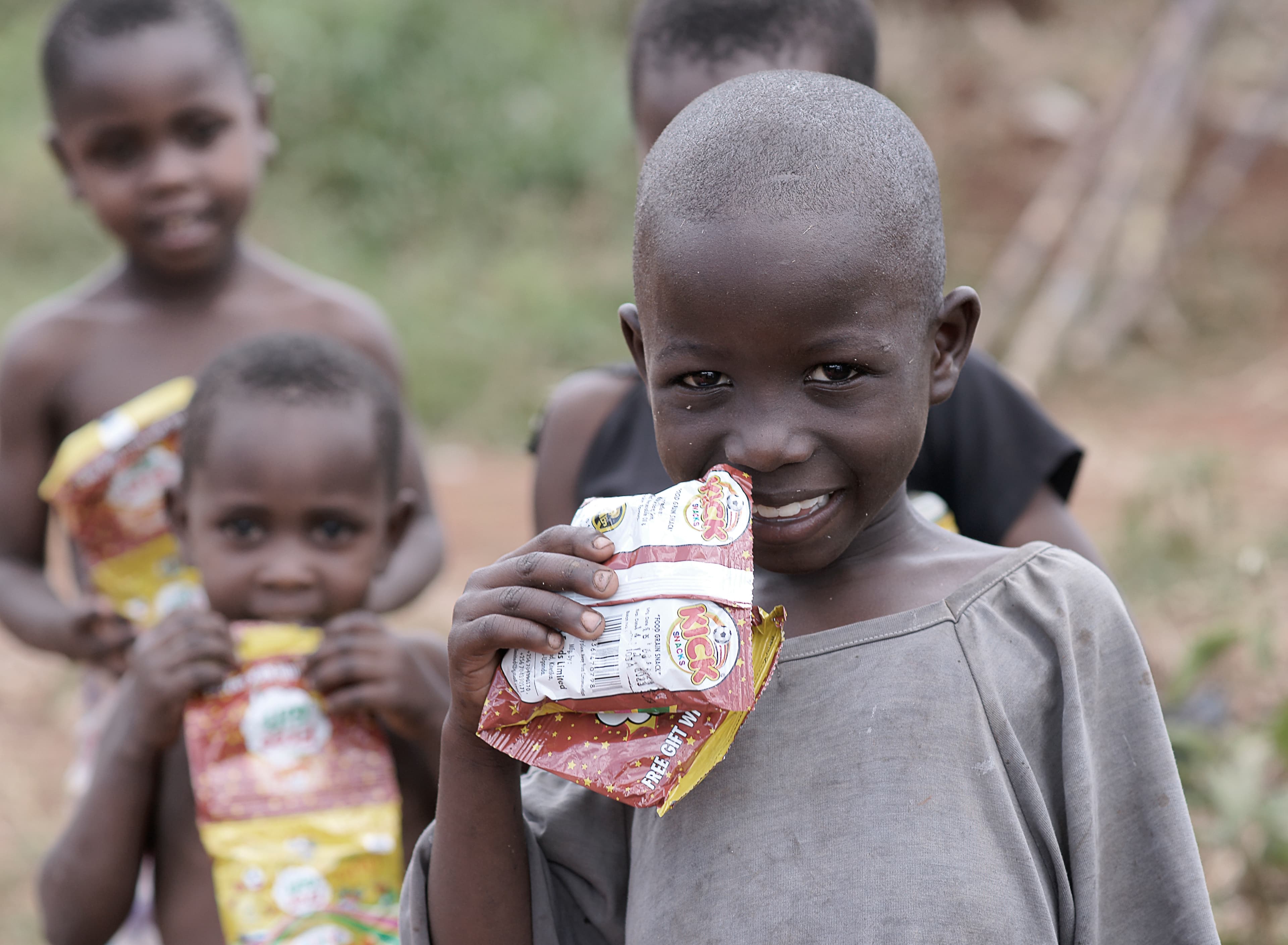 Two children standing close together outdoors, one holding a food packet and smiling