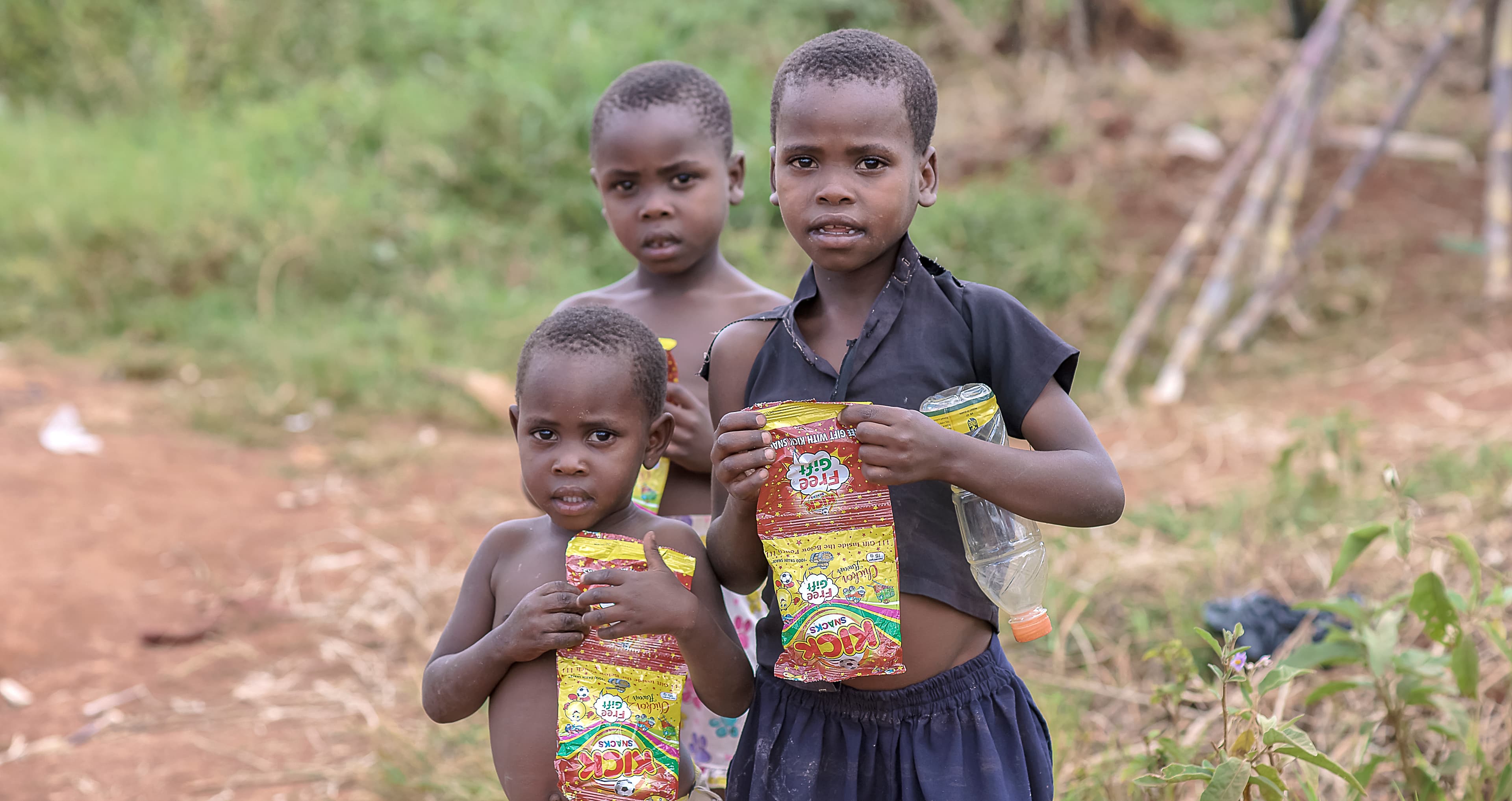 Two young children standing together outdoors, one holding a food packet
