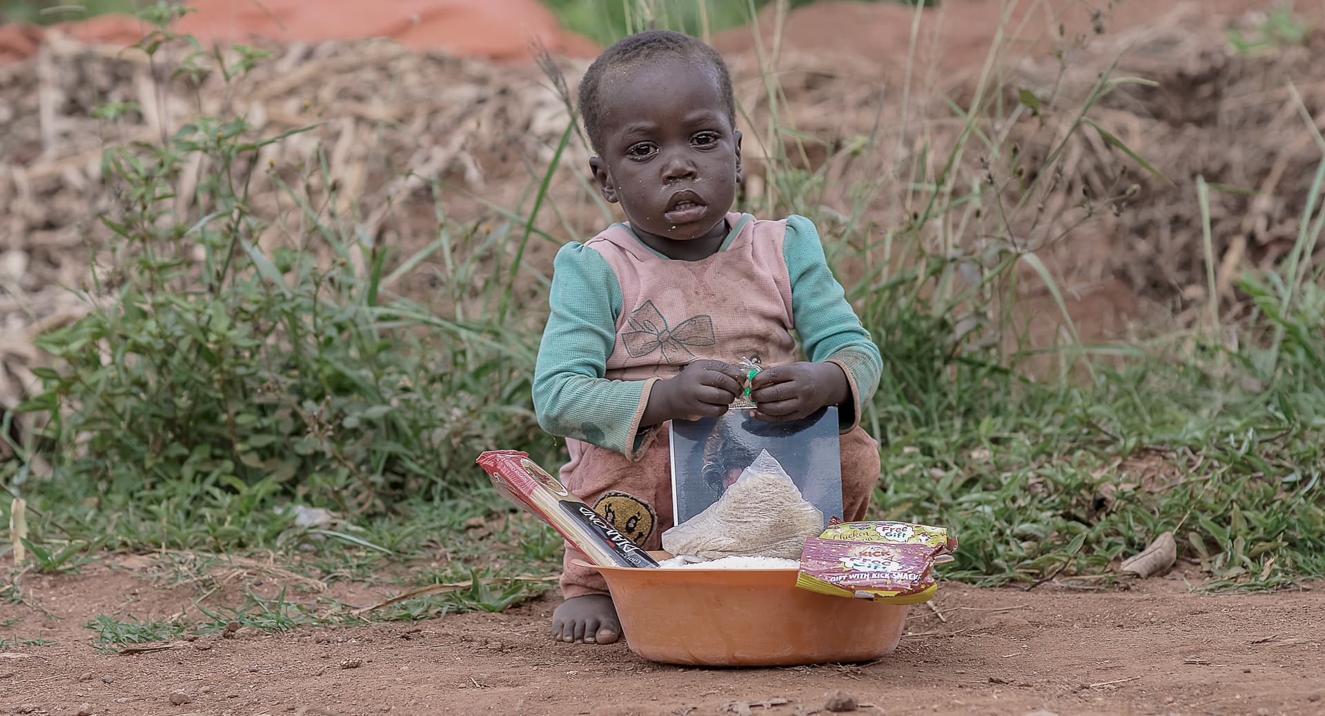 Small child sitting outdoors eating from a bowl placed on the ground