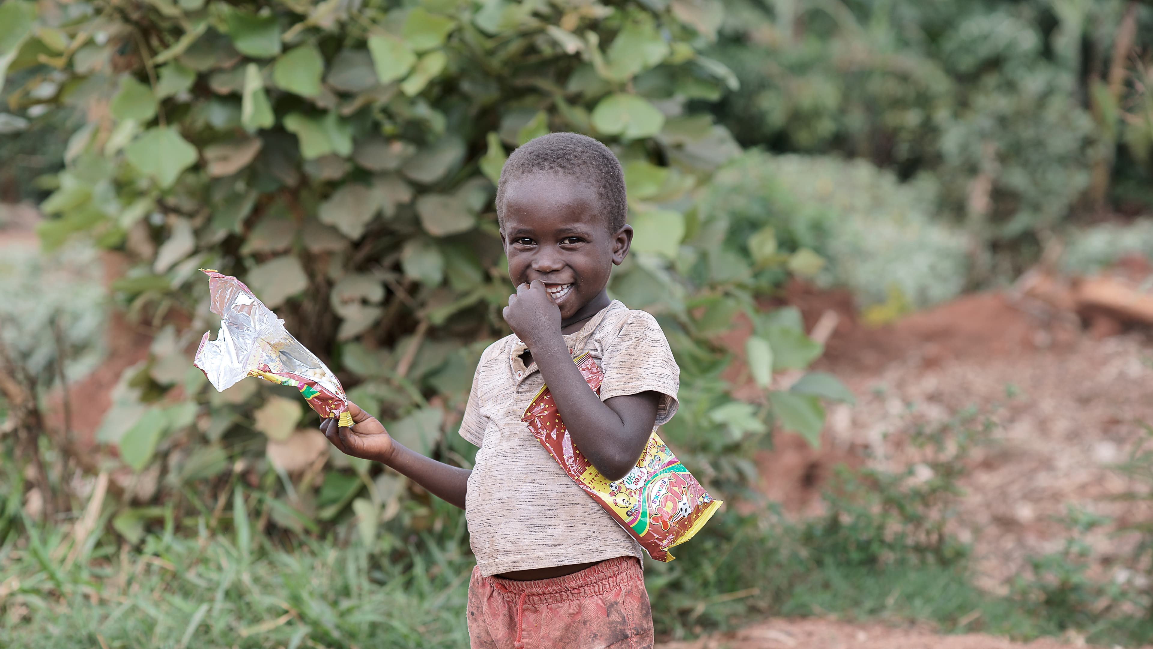 Child standing among green bushes holding a food packet and smiling
