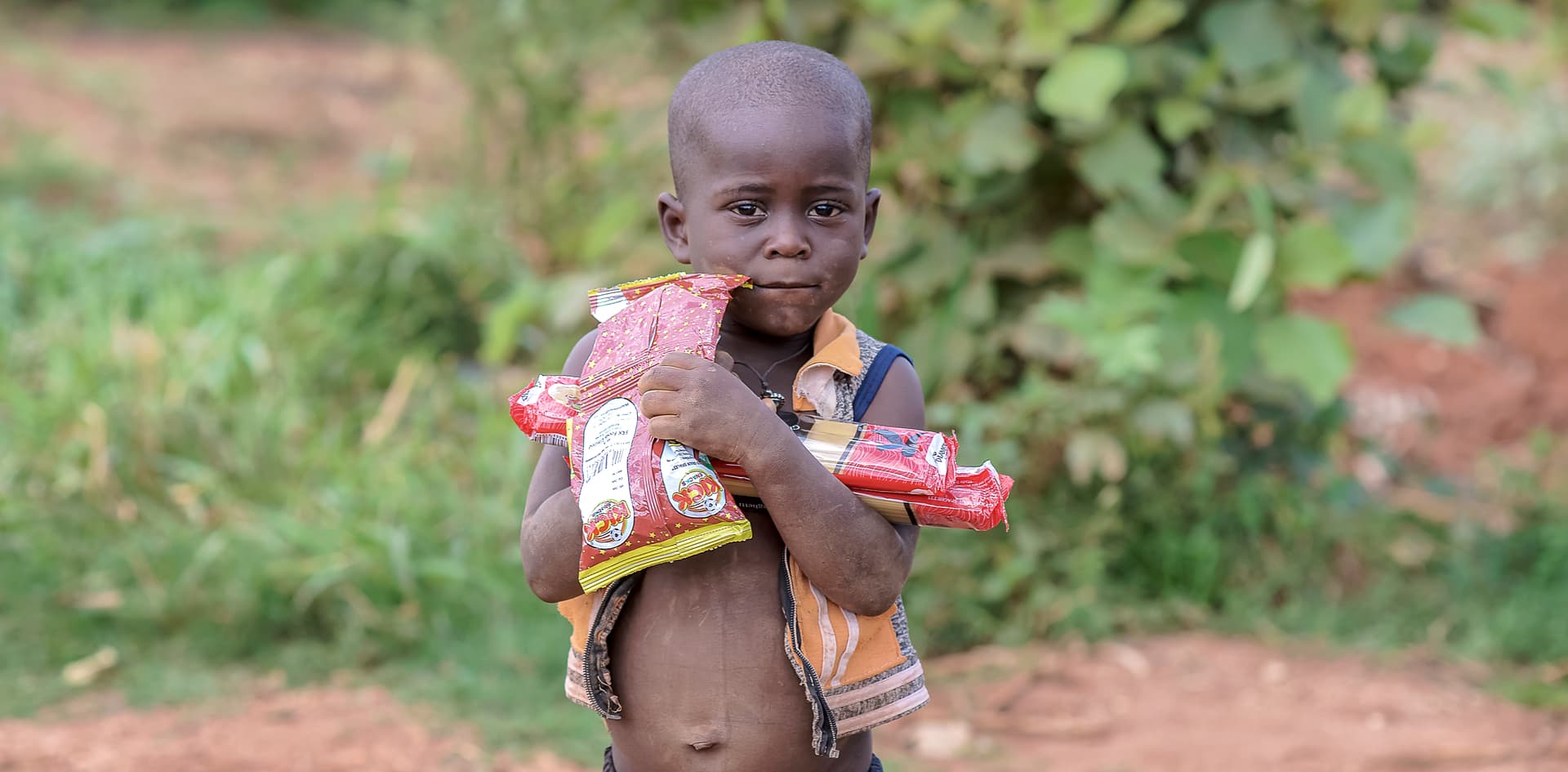 Child in a green top holding food packets outdoors among tall grass
