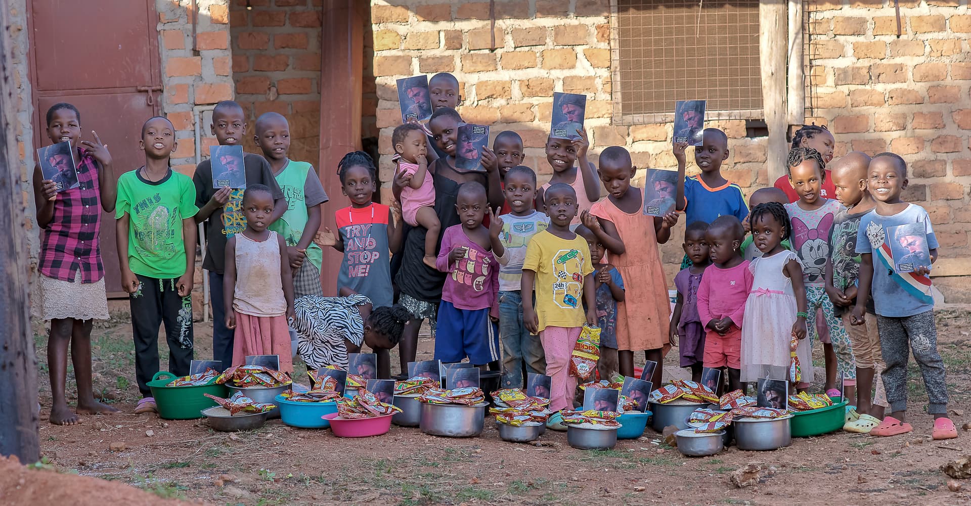 Line of children standing against a brick wall with food bowls set on the ground in front of them