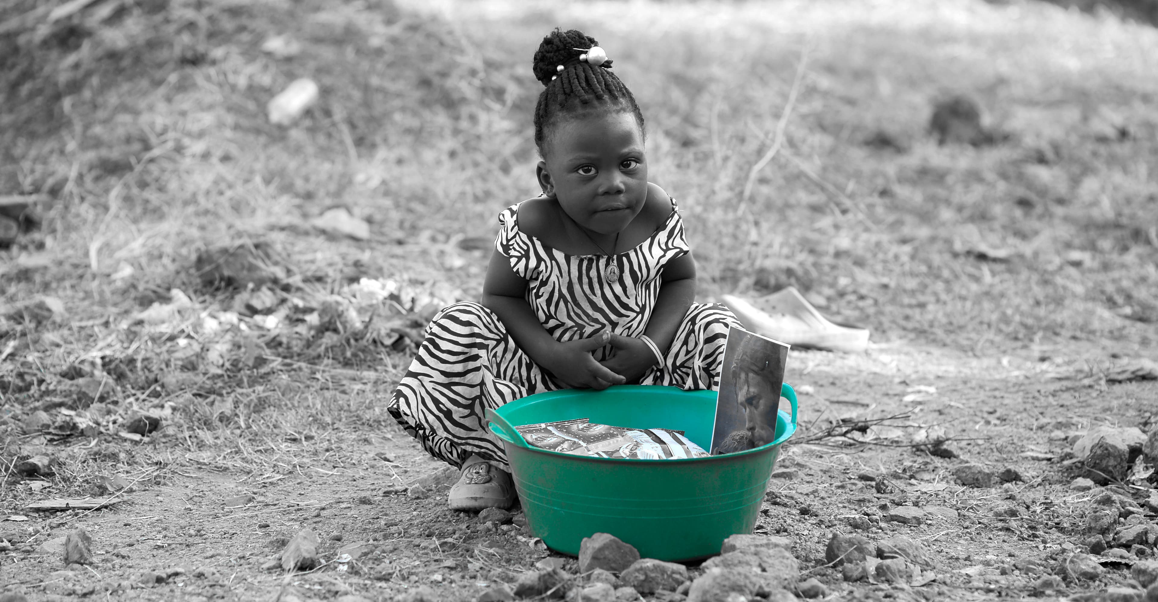 Black and white photo of a young girl sitting on the ground beside a green basin