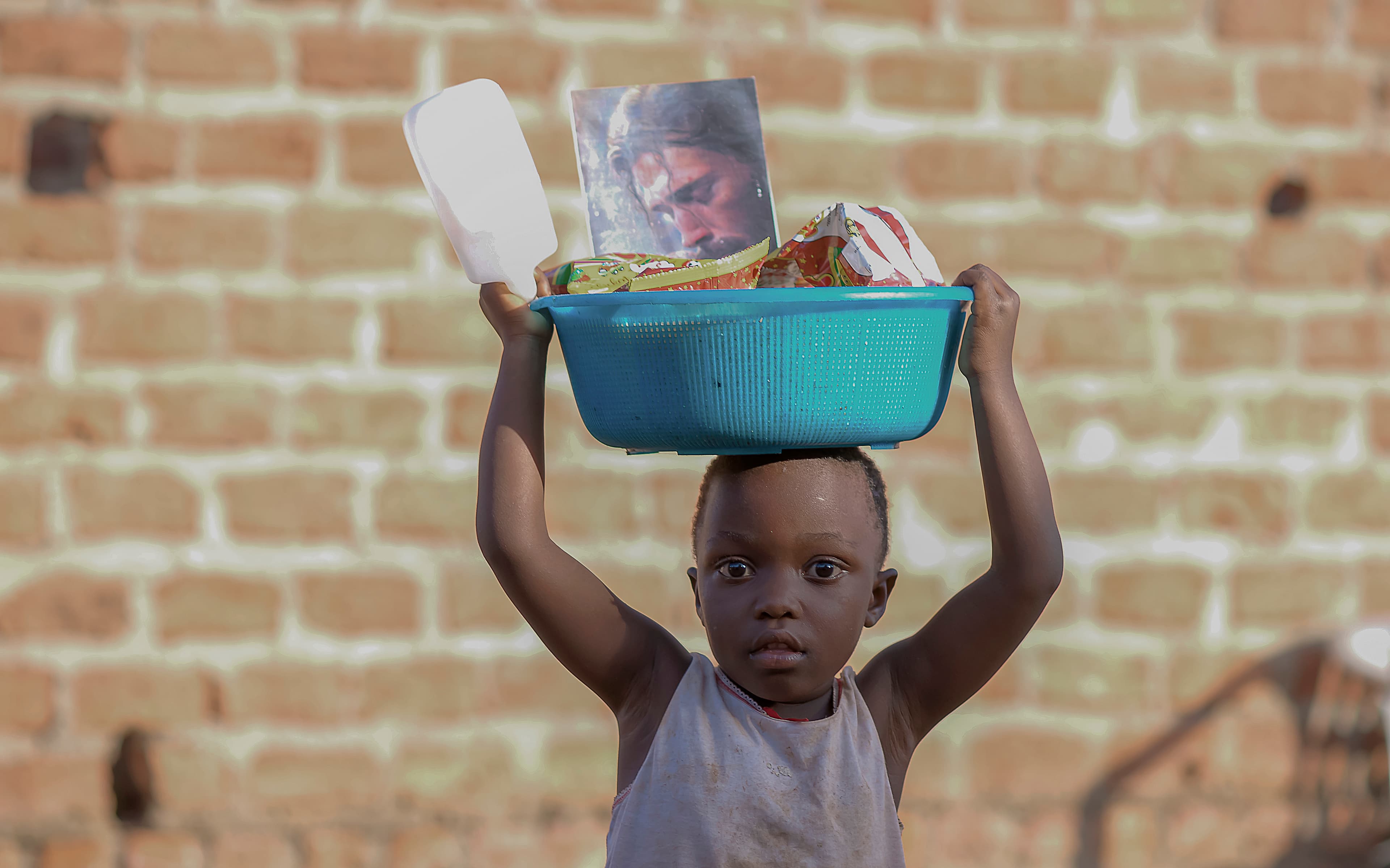 Black and white image of a small child carrying a light-colored basin of food on their head