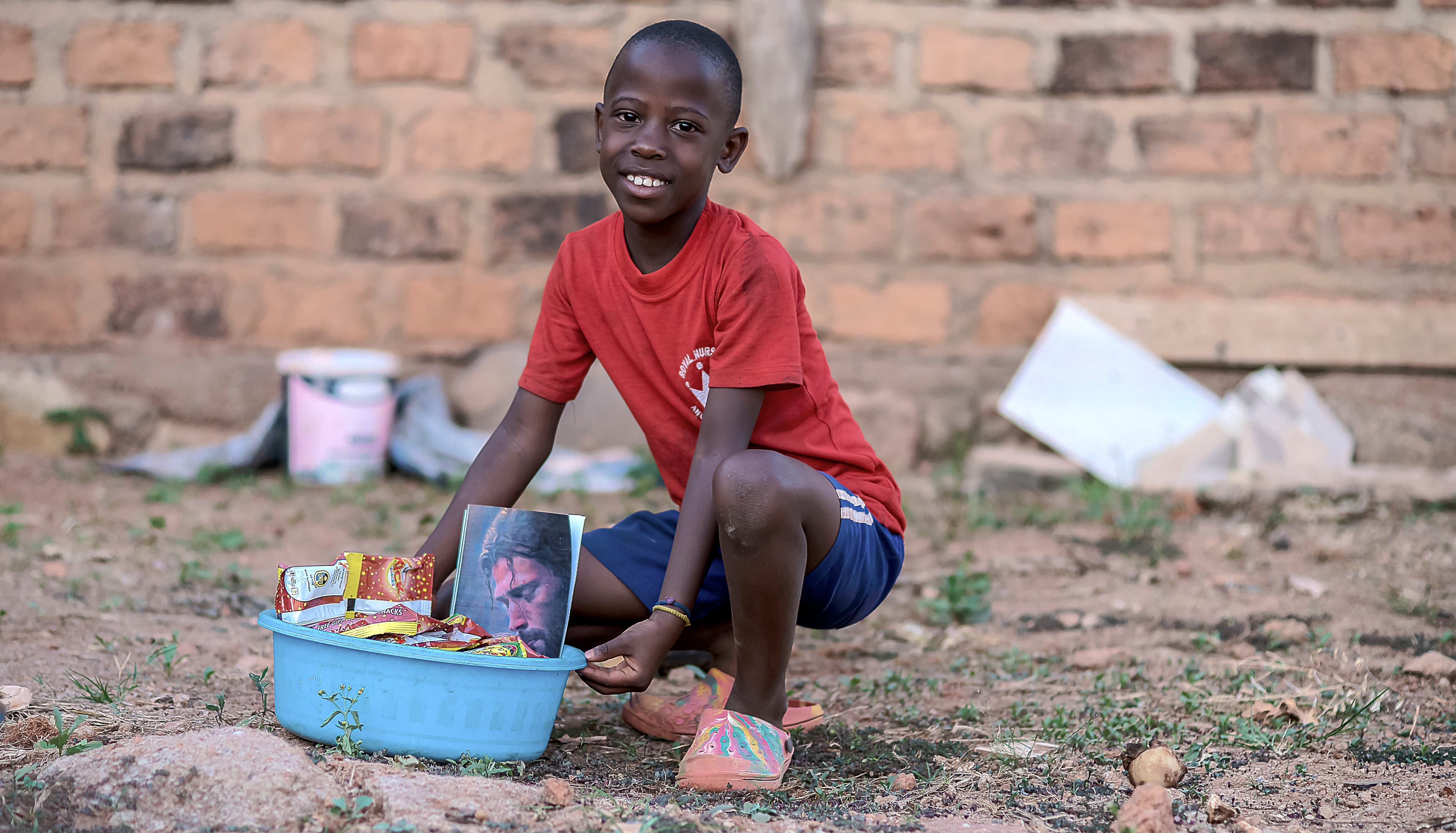 Smiling boy crouched beside a blue basin of food packets near a brick wall