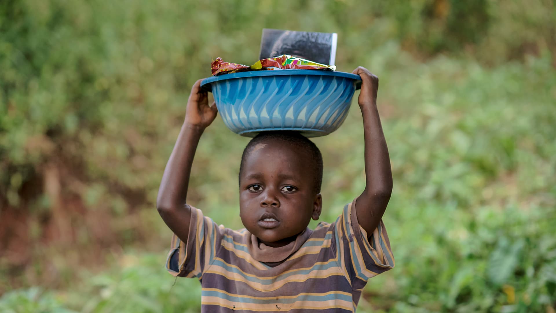 Boy carries a blue basin of food on his head while walking through greenery.