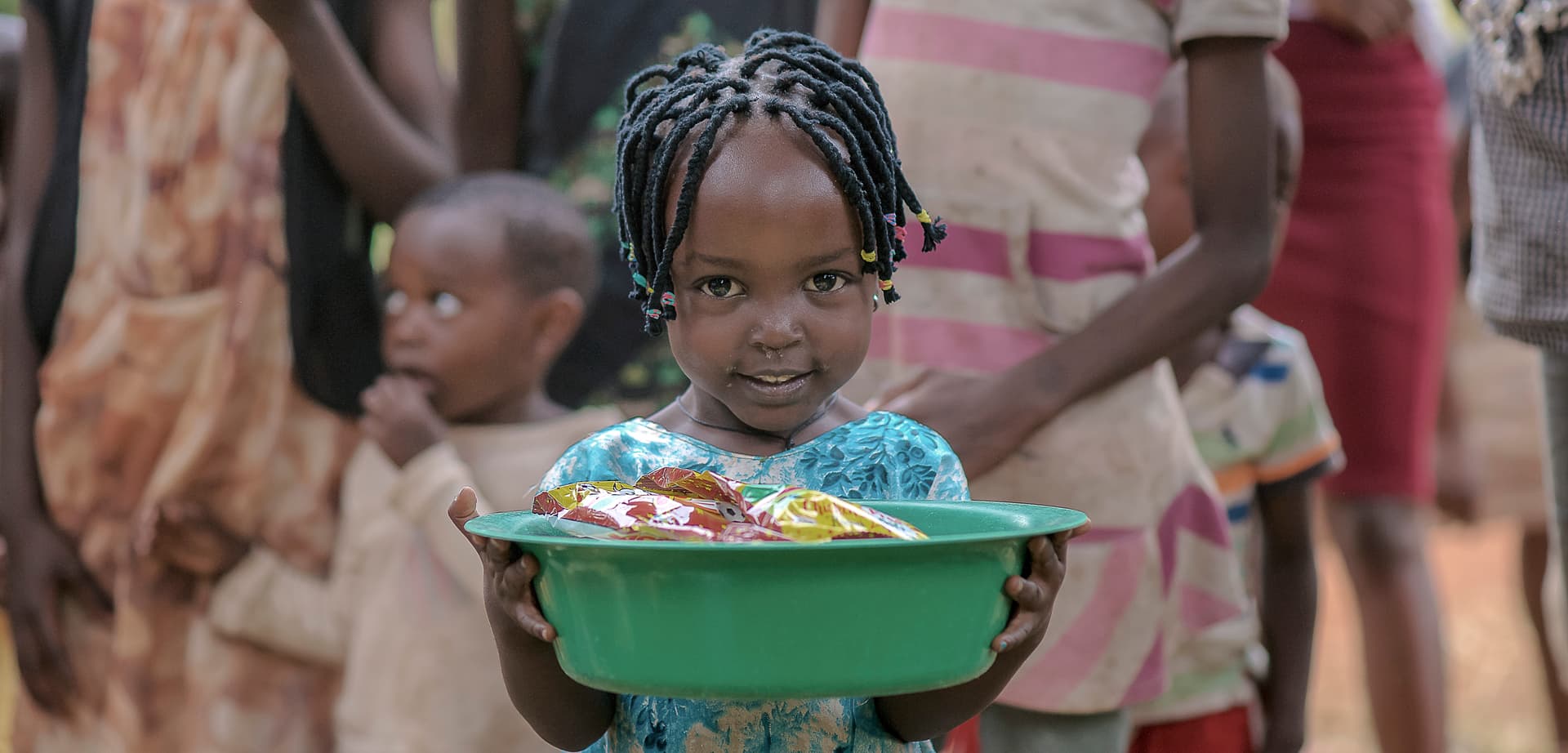 Two girls carry a green basin of food items while other children stand nearby.