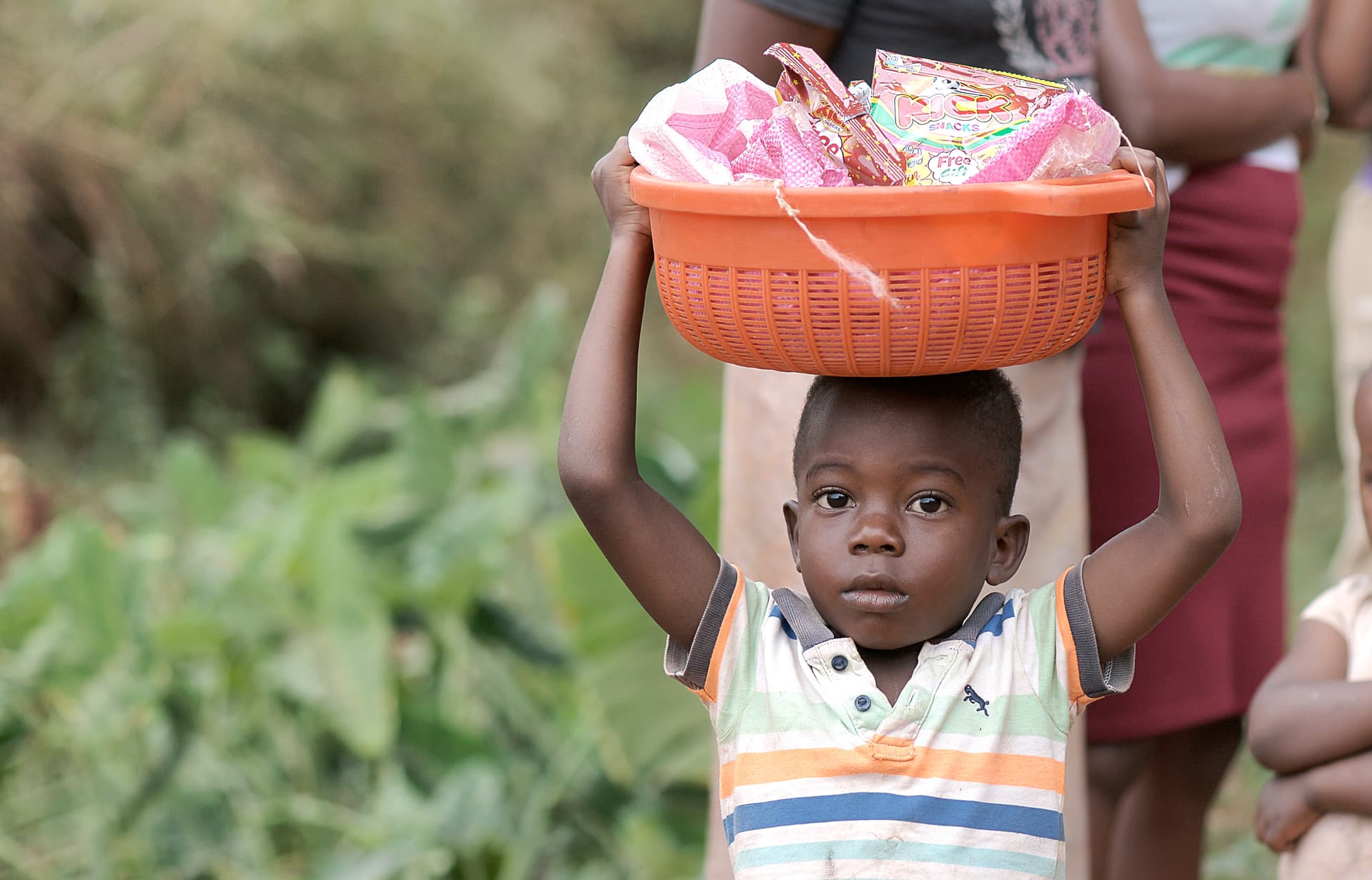 Boy carries an orange basket of food on his head while walking through a field.