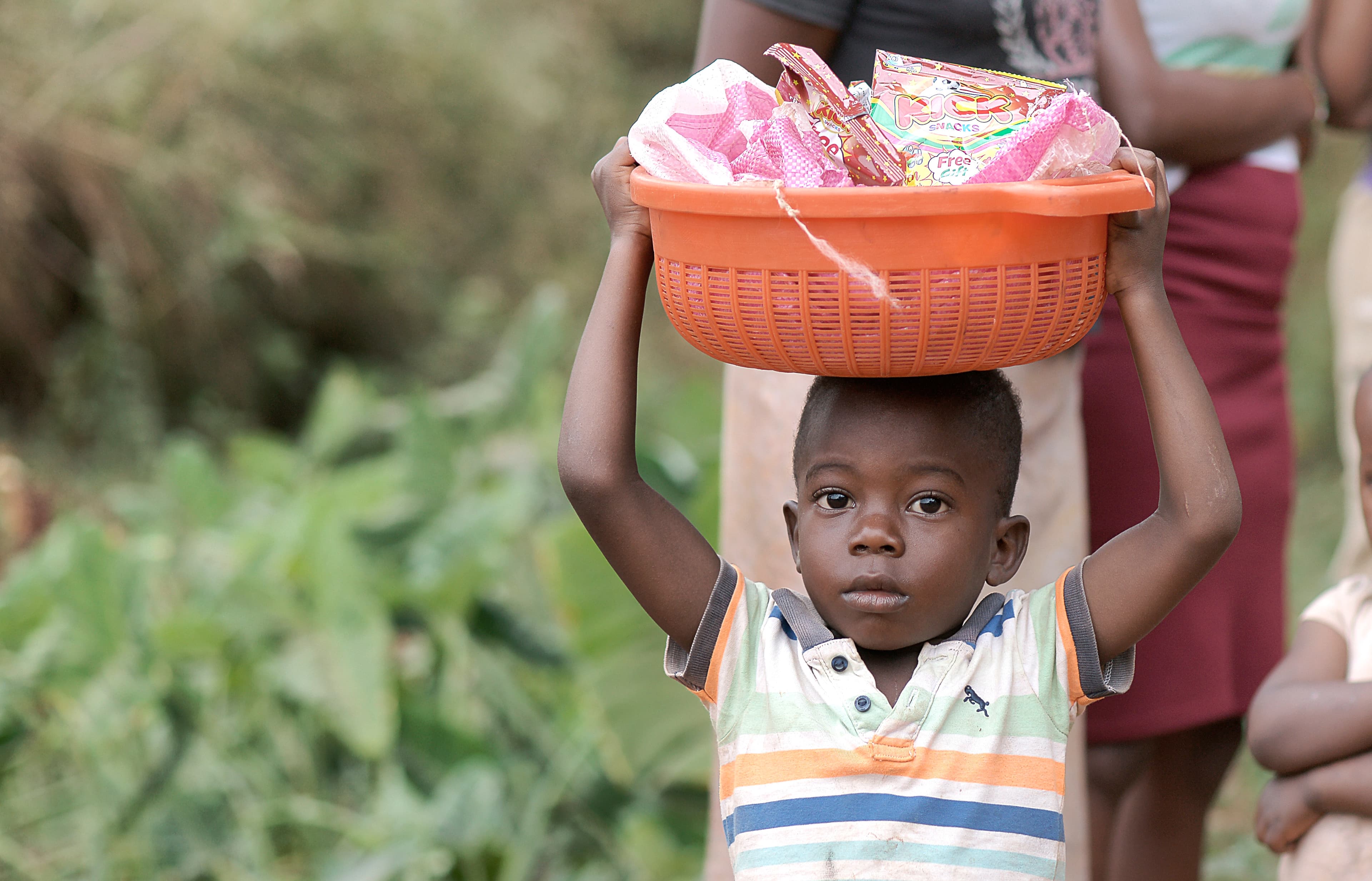Boy carries an orange basket of food on his head while walking through a field.
