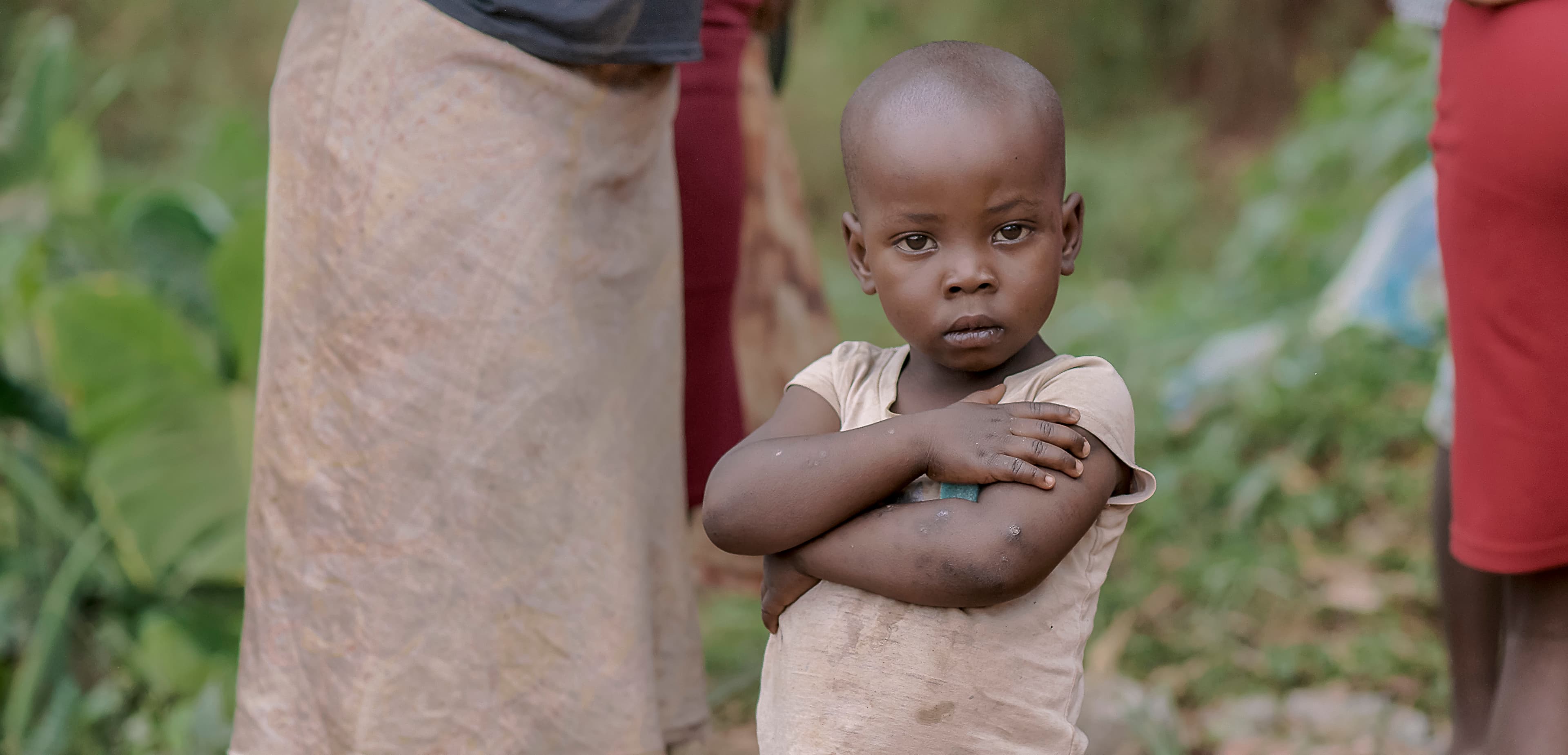 Woman with arms crossed carries a young child on her hip outdoors.