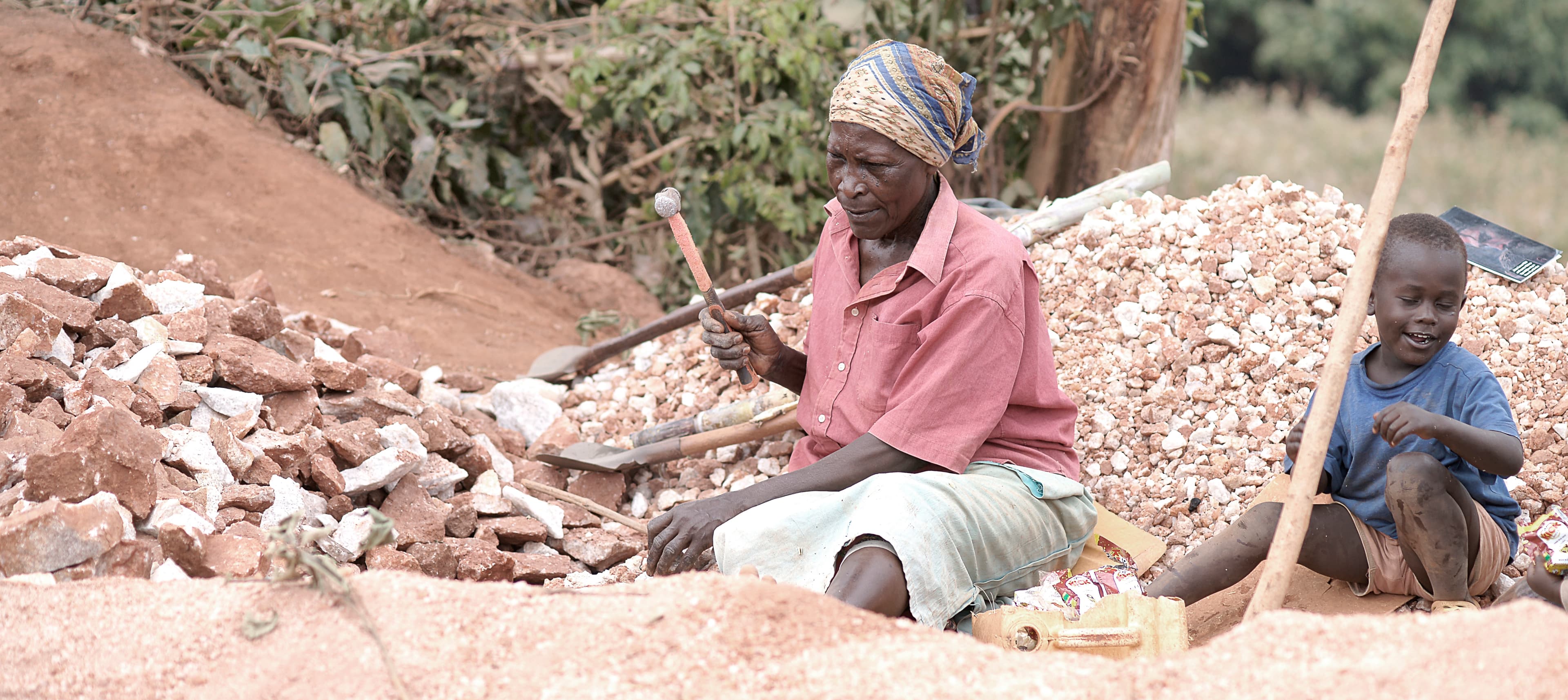 Woman and young child sit among rock piles with hammers at a quarry site.
