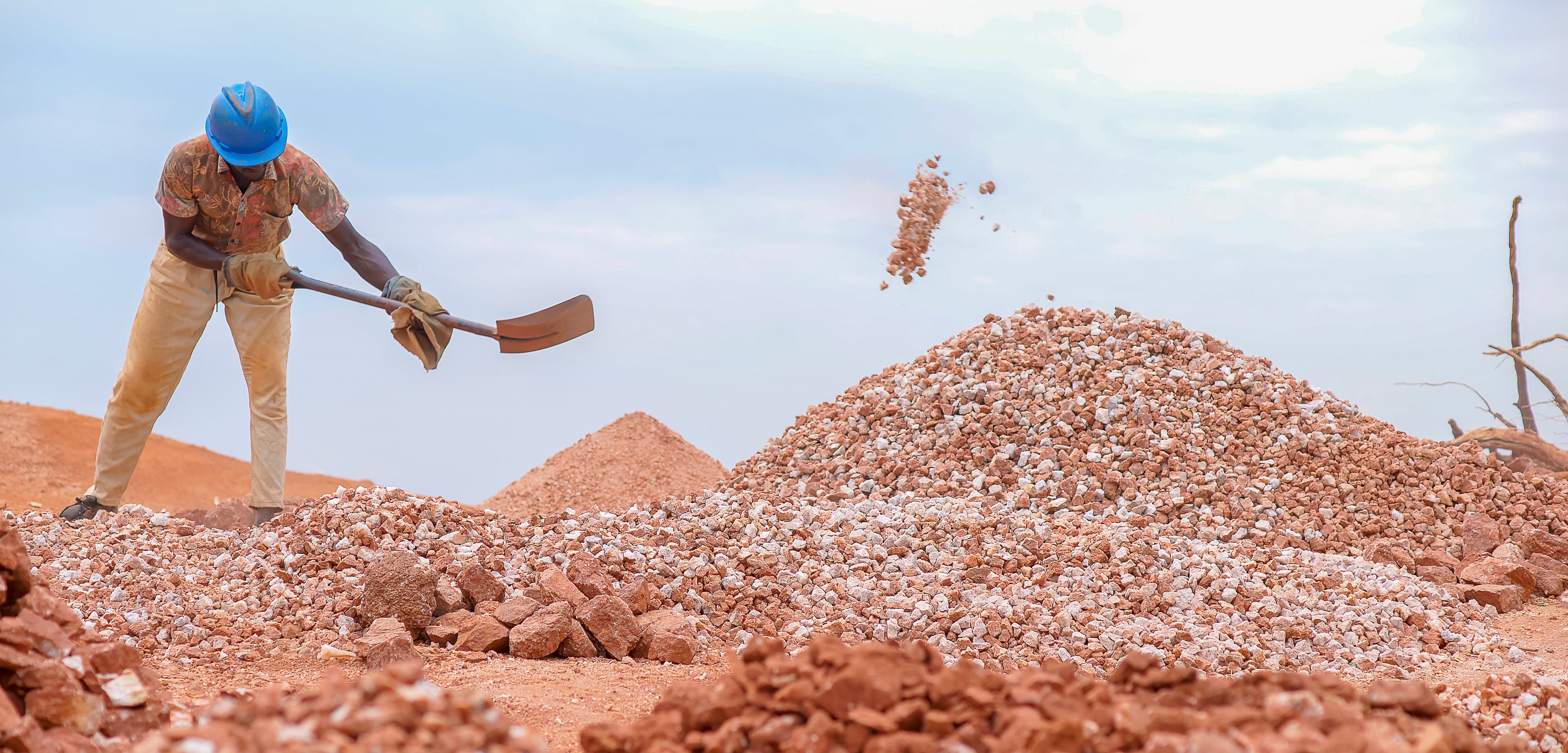 Man in a hard hat shovels crushed rock at a quarry site as another worker stands atop a pile.