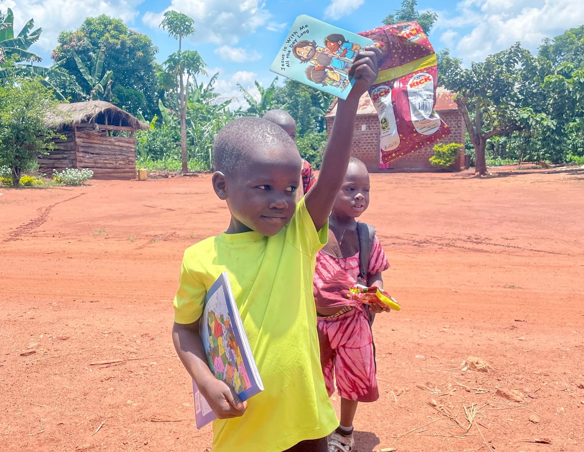 Young boy in a yellow shirt holds up an illustrated picture book near a hut.