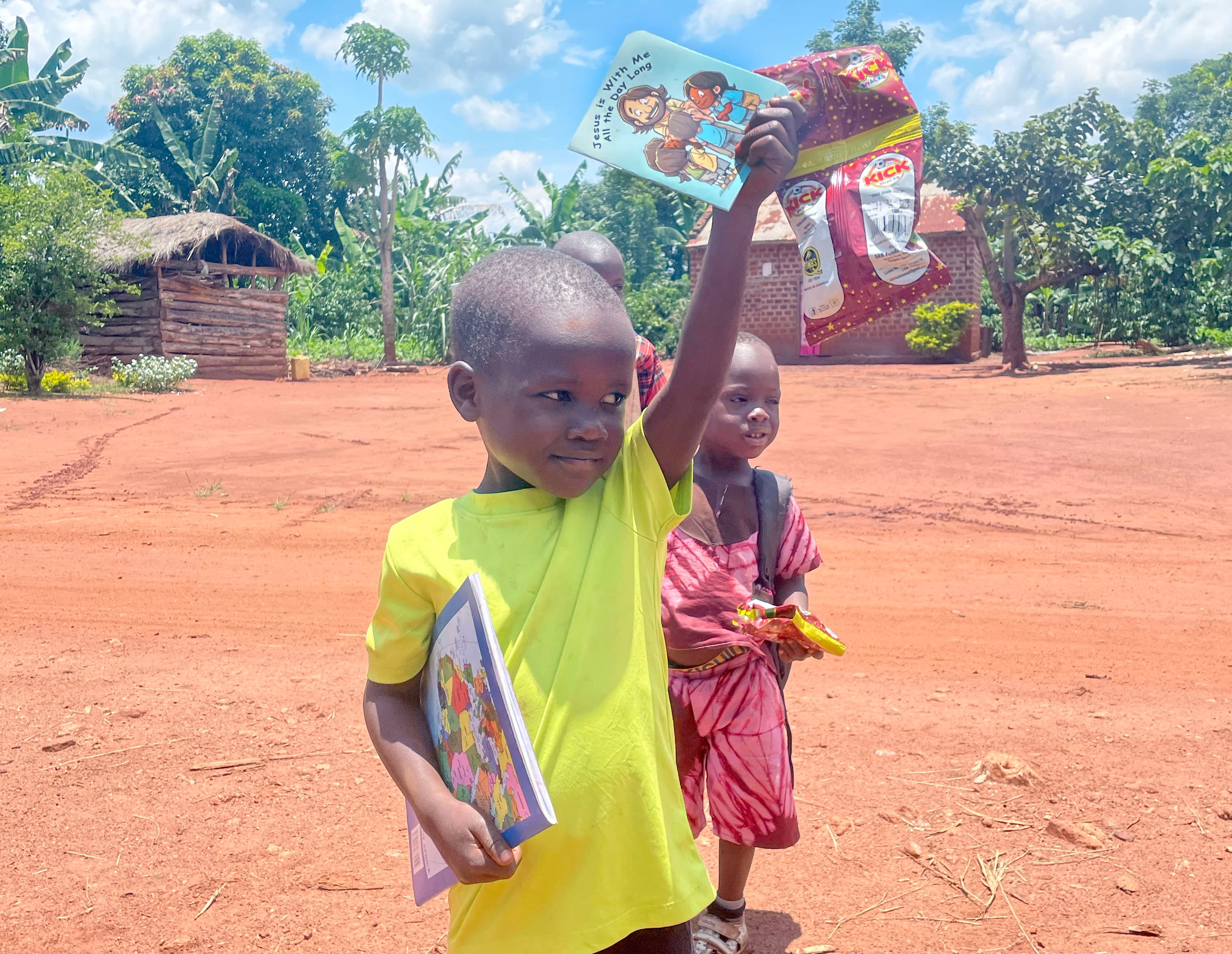 Young boy in a yellow shirt holds up an illustrated picture book near a hut.