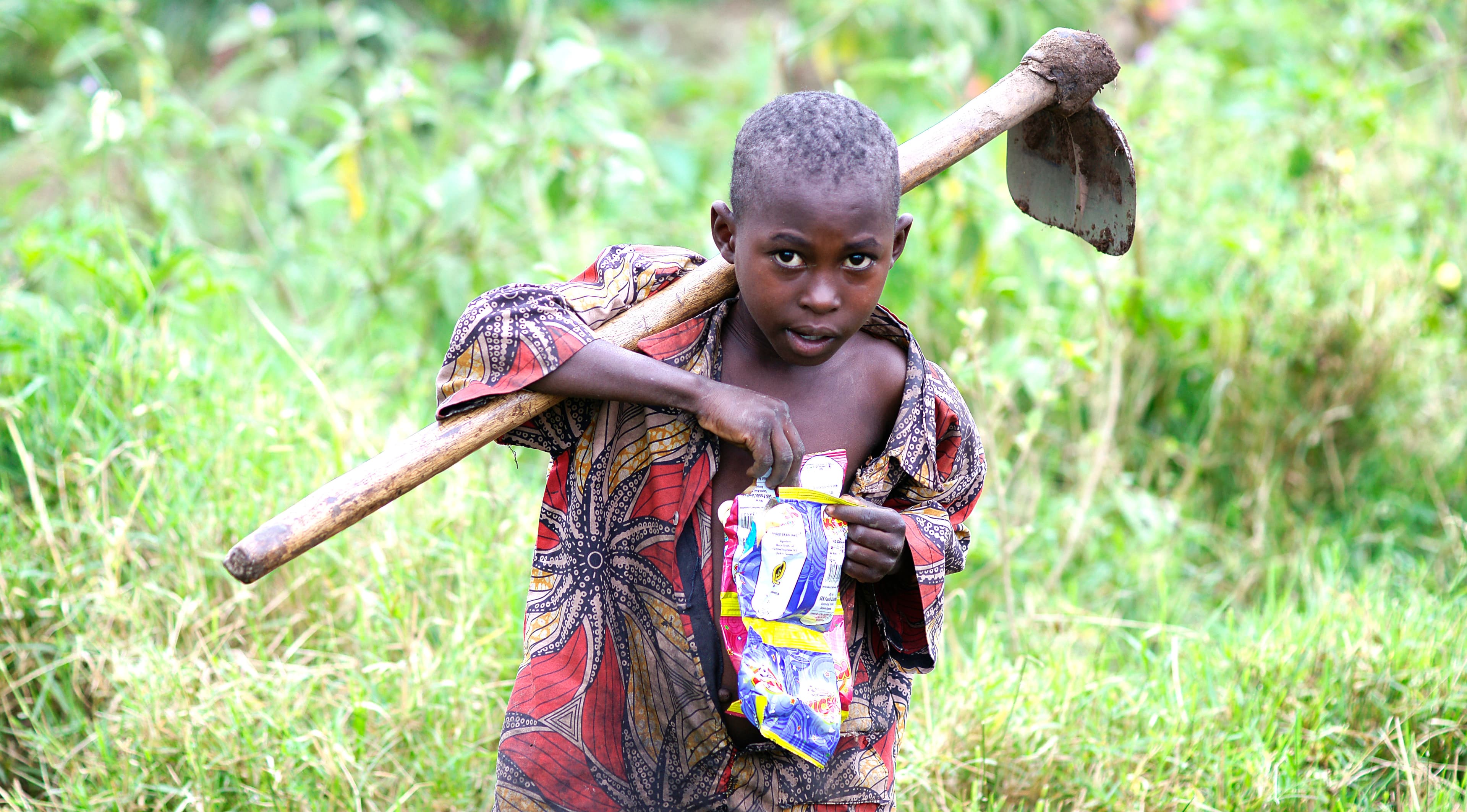 Girl in a colorful dress carries a hoe on her shoulder while walking through a field.