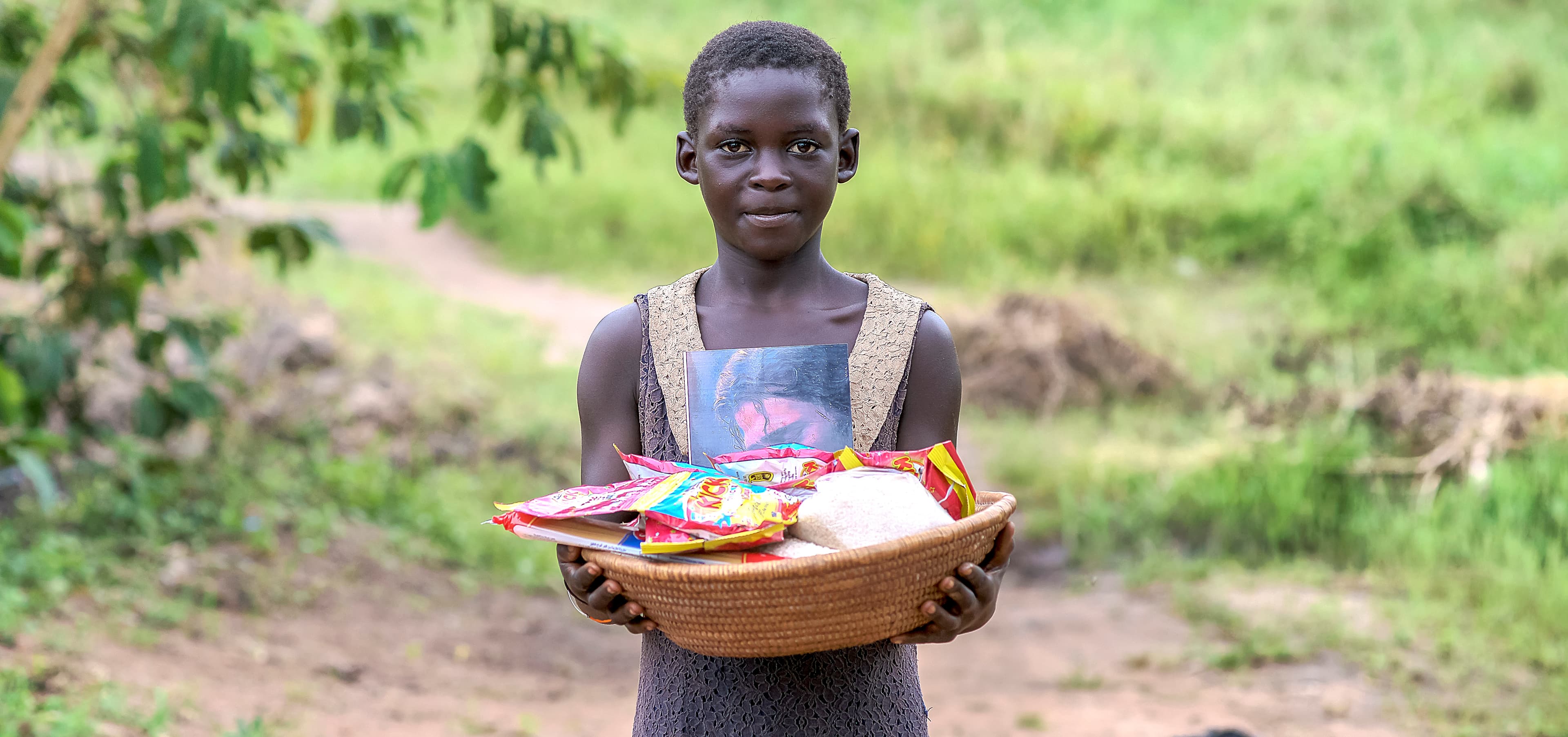 Girl carries a woven basket of food items on her head outdoors.