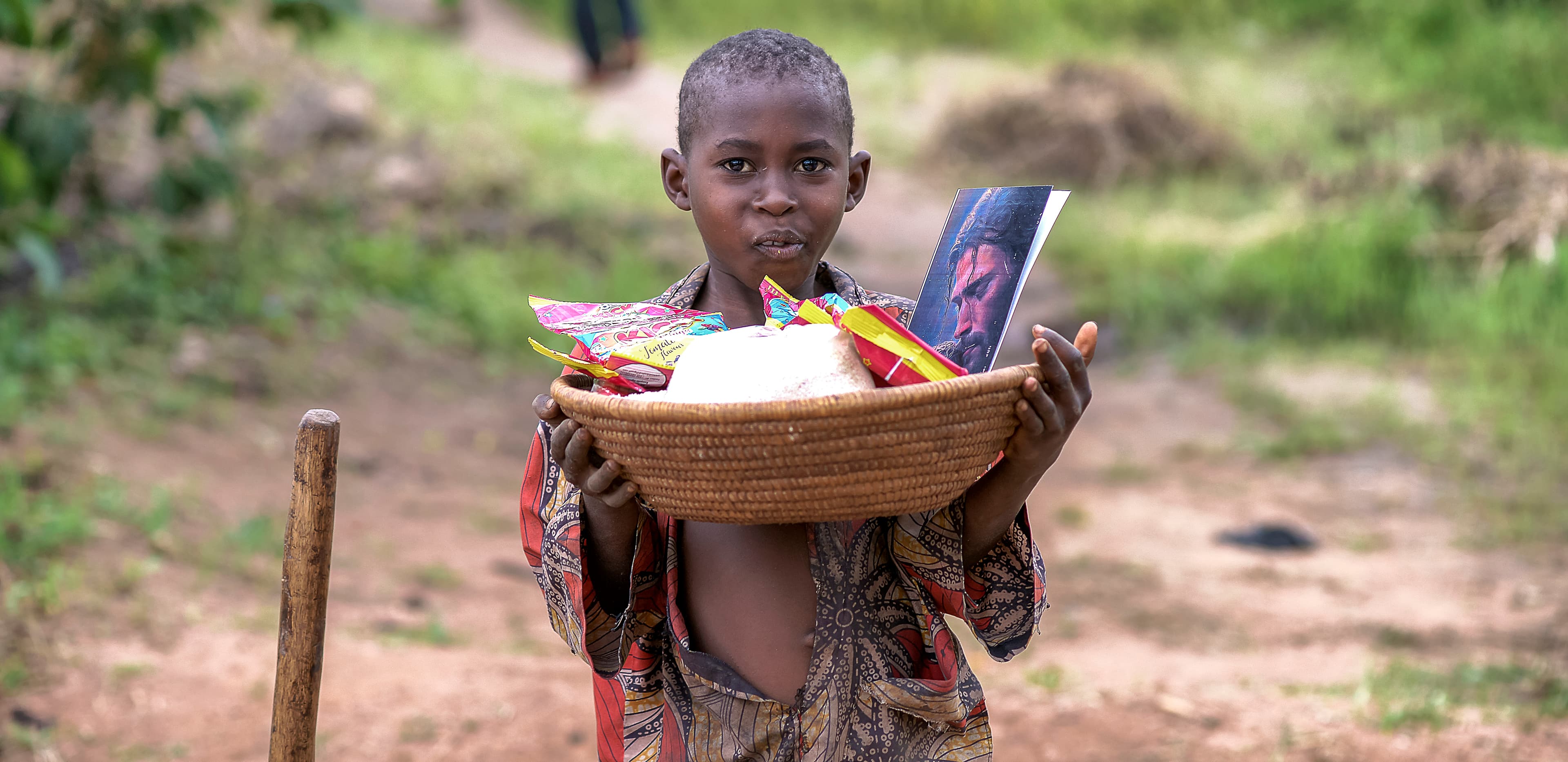 A boy stands on a dirt path holding a basin of gospel tracts and food items.
