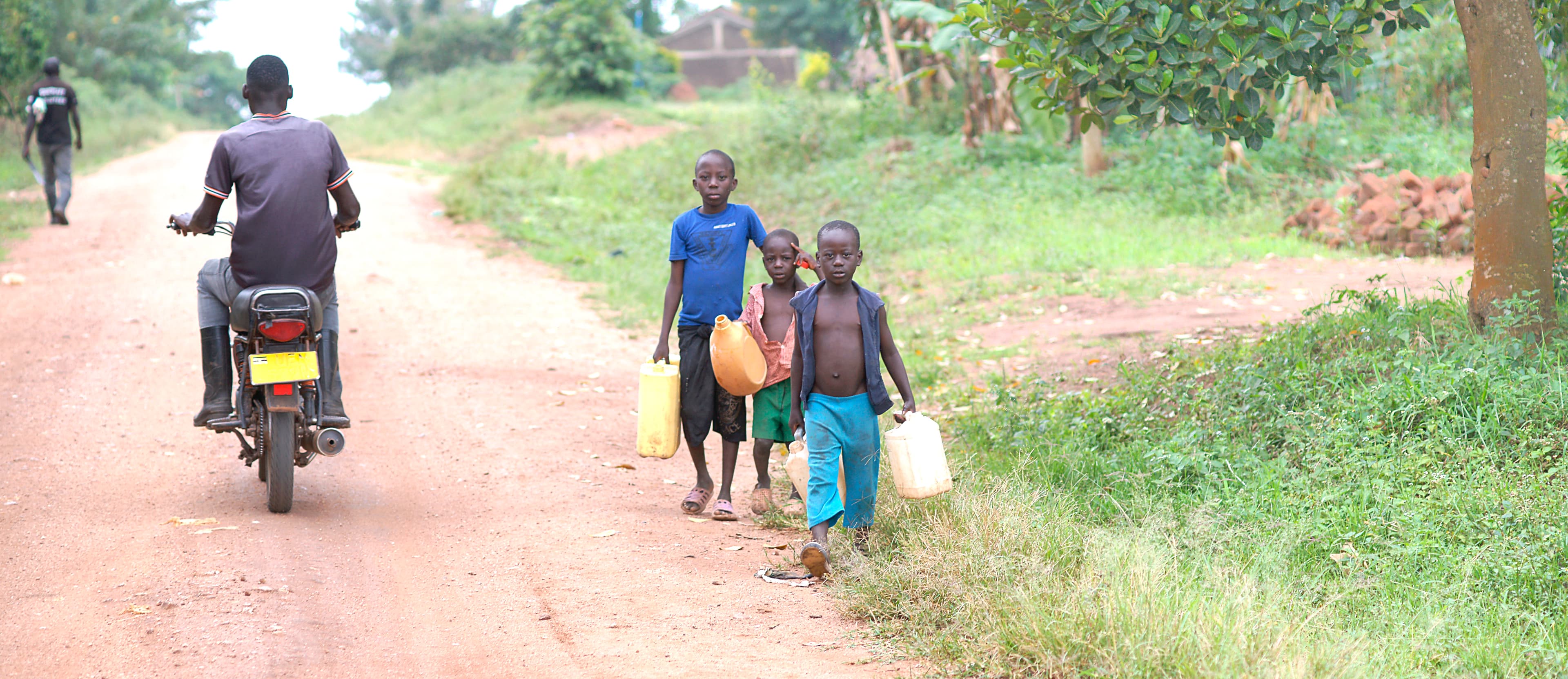 A man rides a motorcycle on a dirt road as children walk nearby.