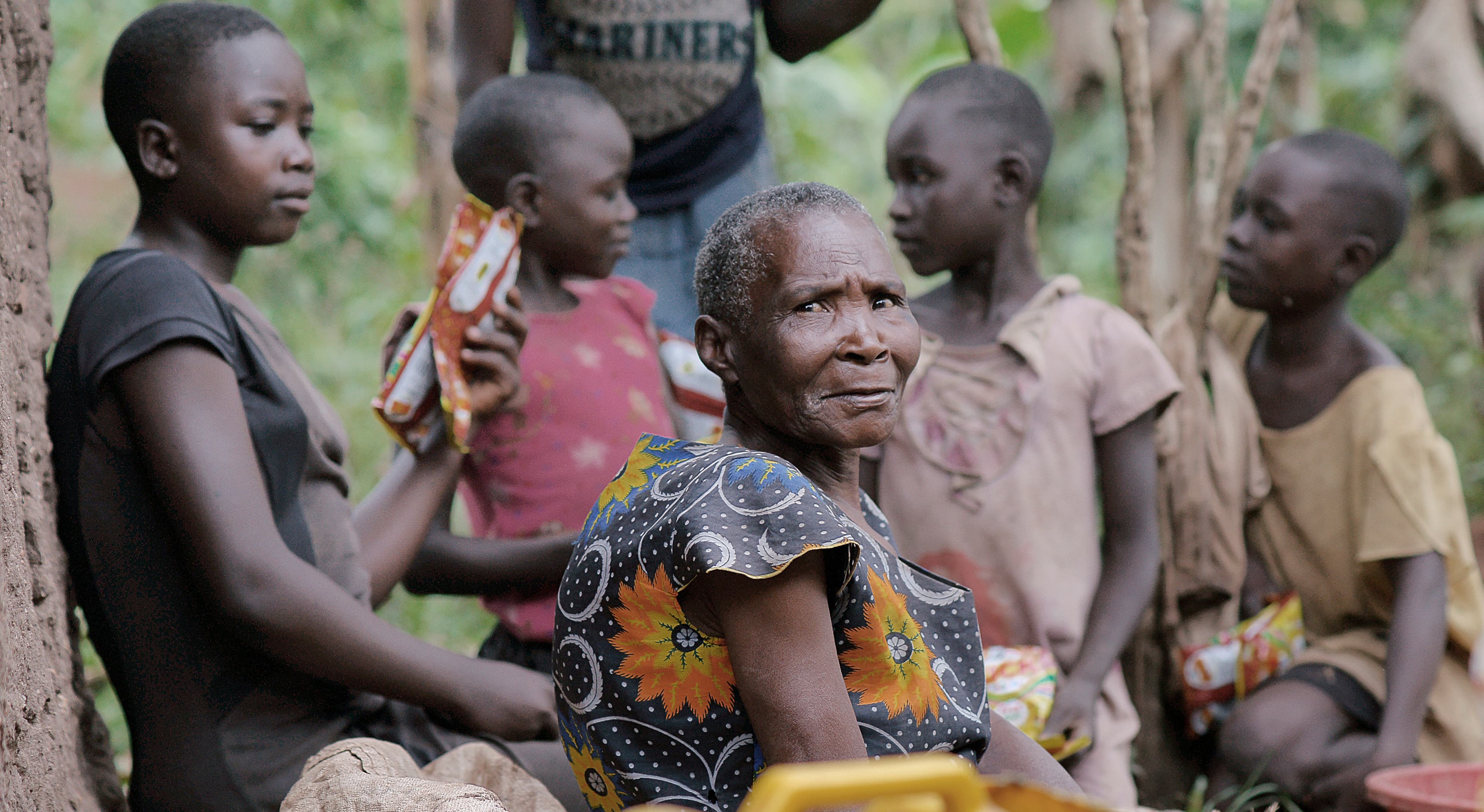 Elderly woman and children gather outdoors holding food and photo items.
