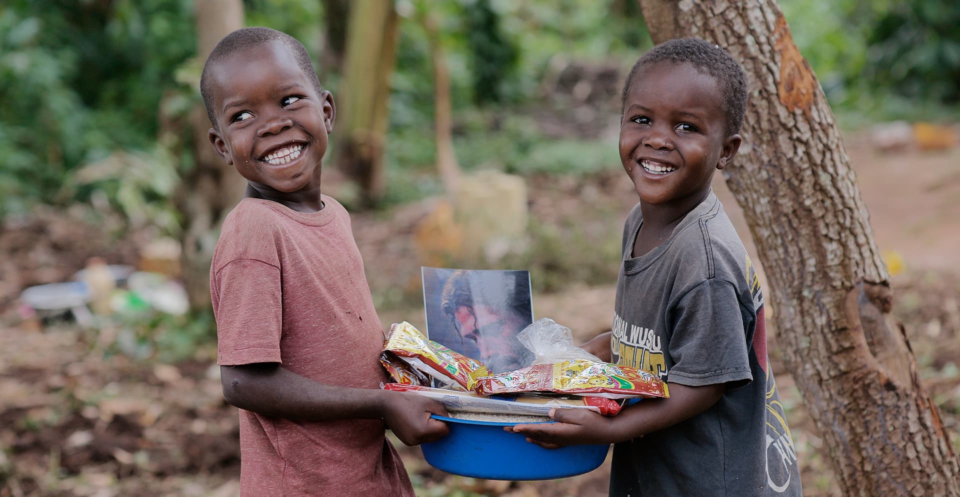 Two boys stand together, one holding a pot of food at his hip.