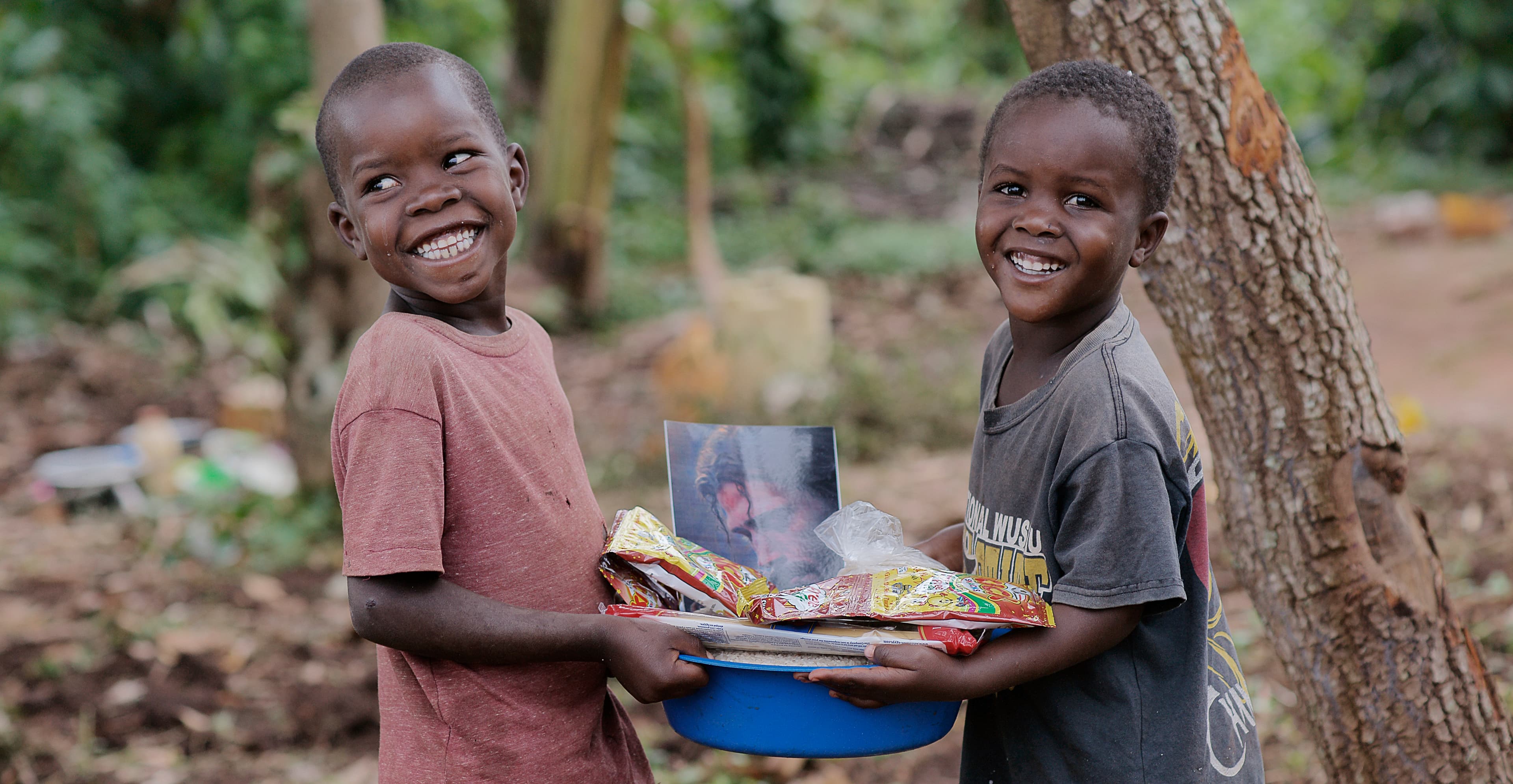 Two boys stand together, one holding a pot of food at his hip.