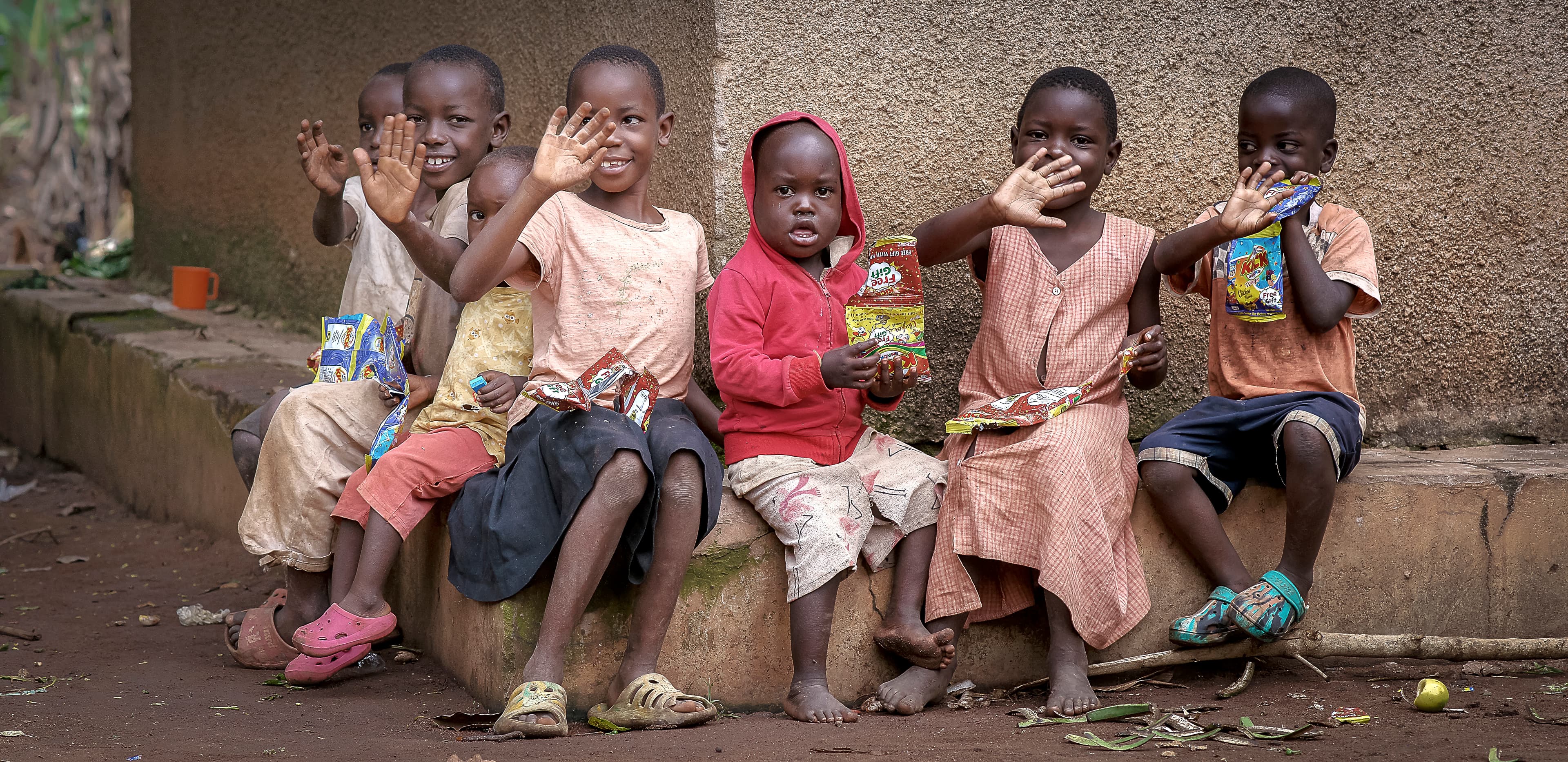 Group of children sit together on a stone step, waving toward the camera.