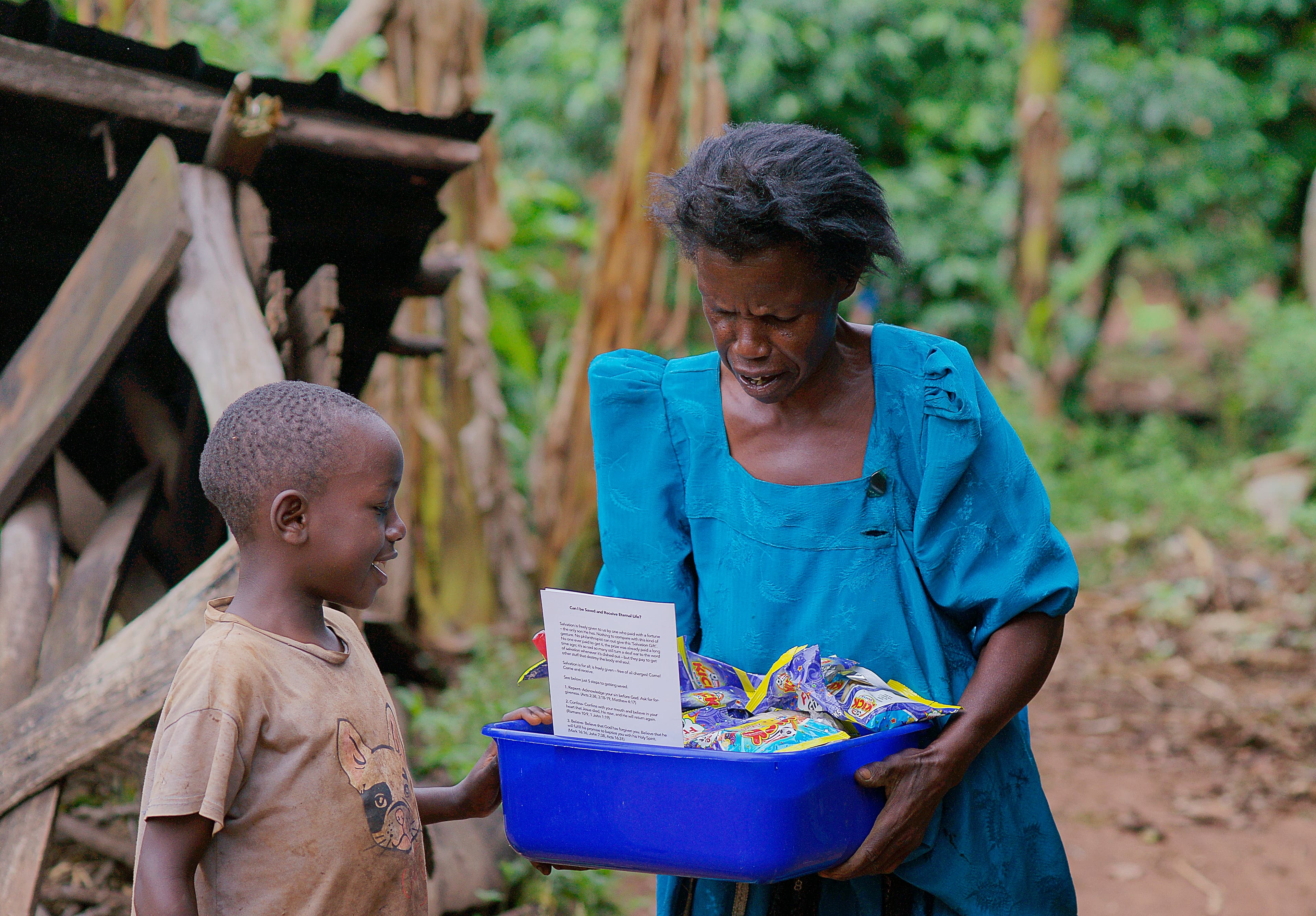 Elderly woman and a boy stand outside a mud-walled house, the boy holding a blue food container.