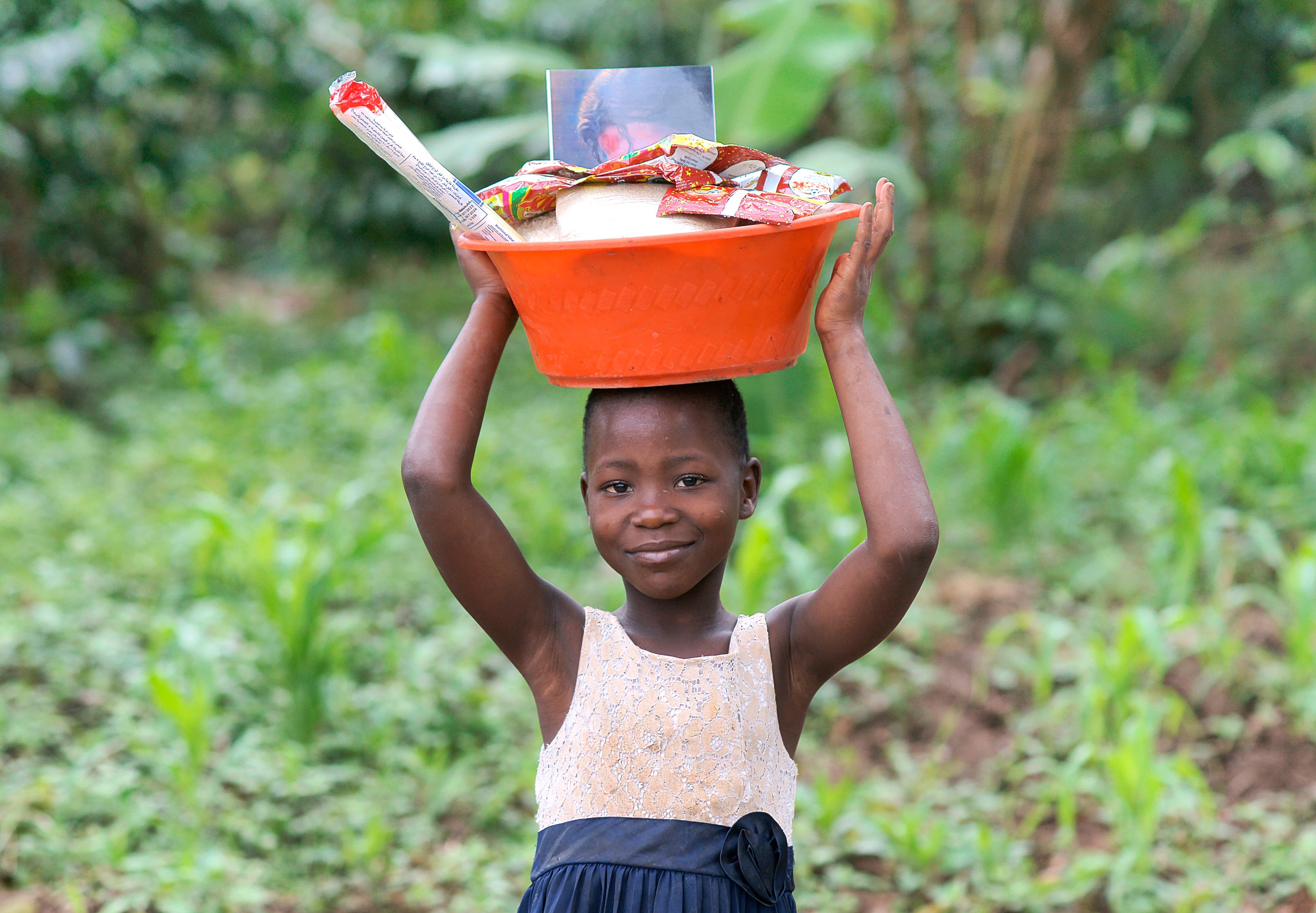 Girl carries an orange basin with food and toiletries balanced on her head in a field.