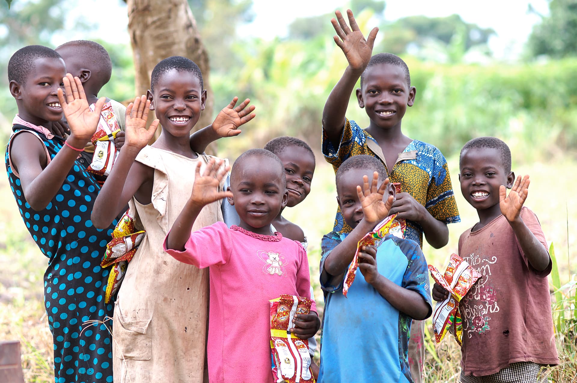 Group of children wave and smile while holding food packages outdoors.