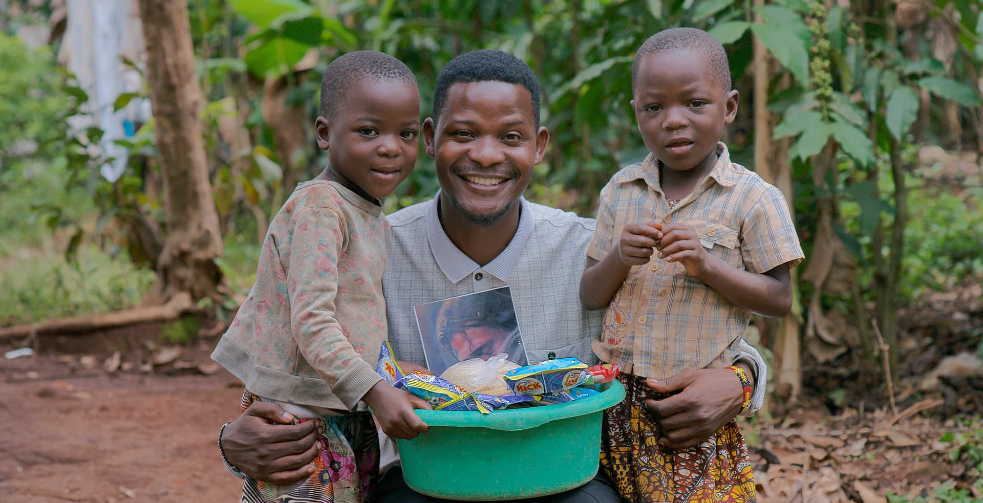 Three children stand together, one holding a teal basin of food and toiletries.