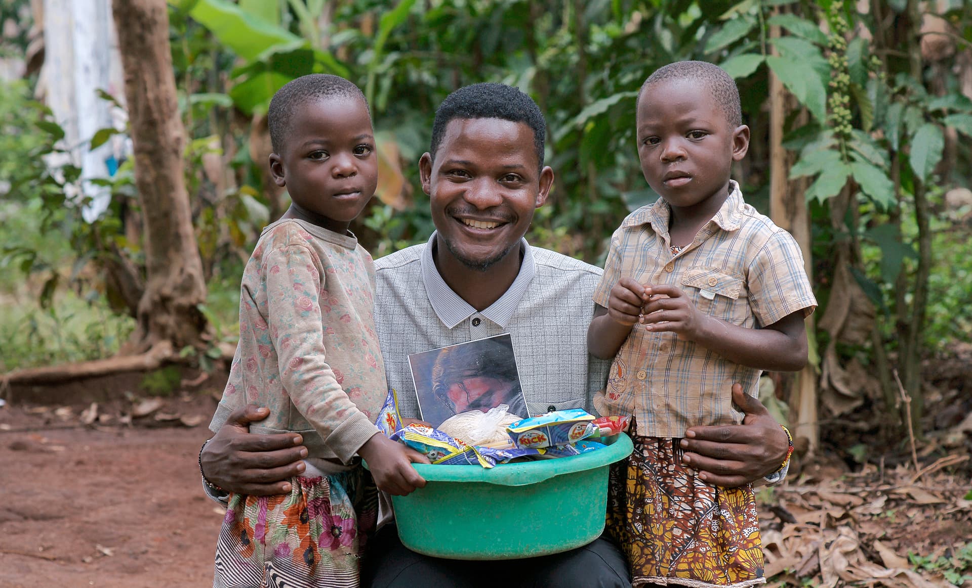 Man stands with two children, one holding a green basin of food items.