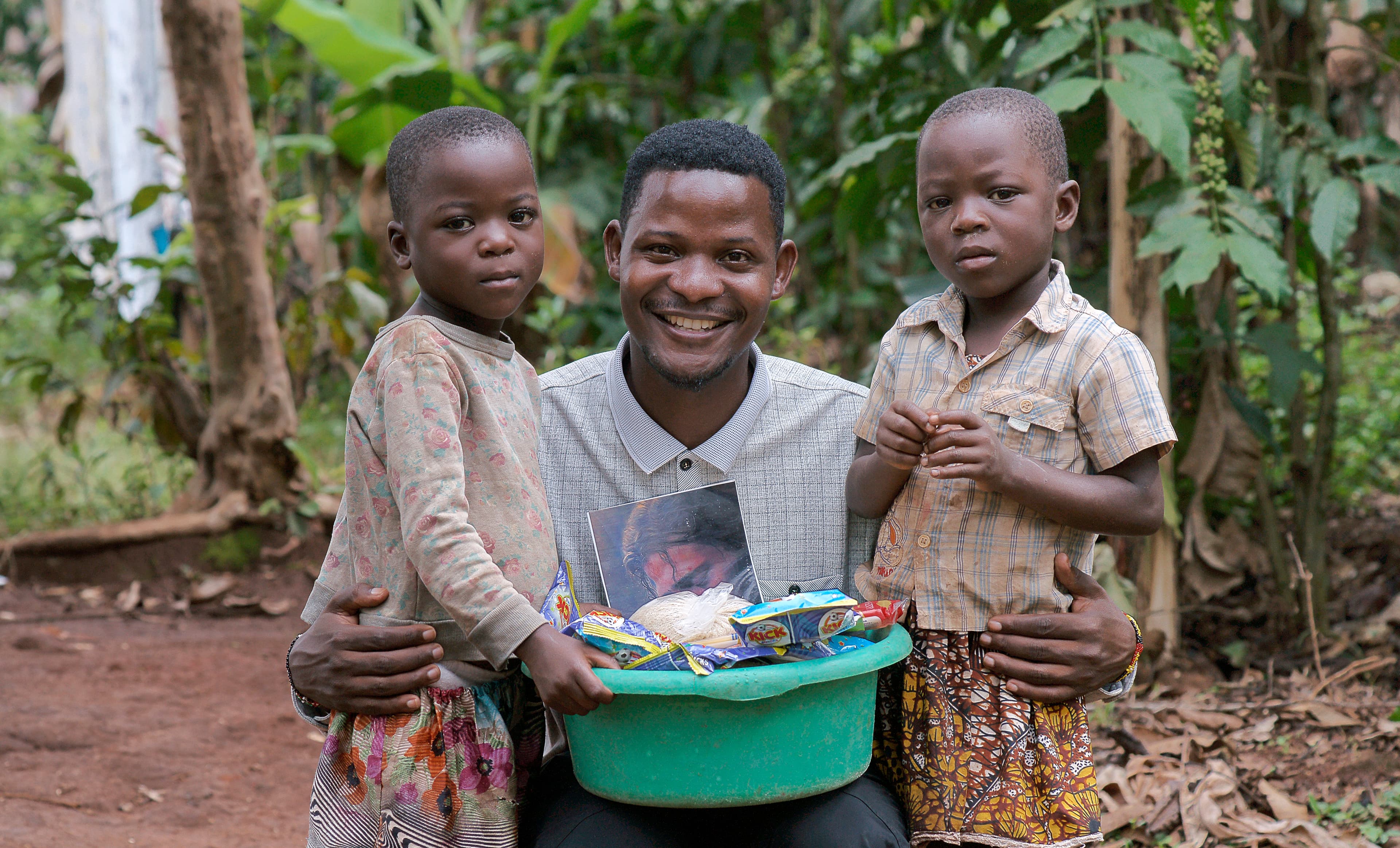 Man stands with two children, one holding a green basin of food items.