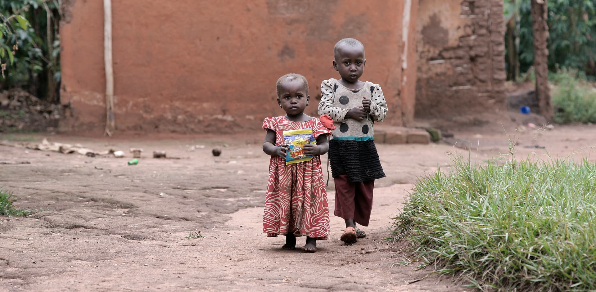 Two women in colorful dresses carry children while walking down a dirt road, seen from a distance.