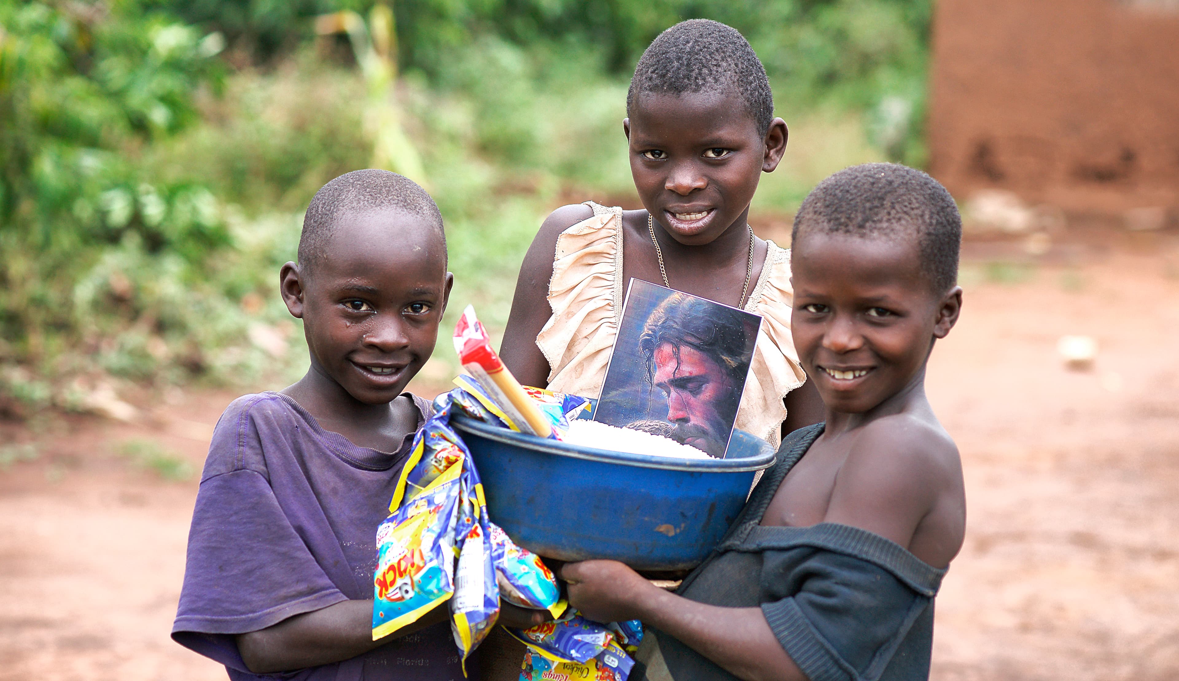 Three children stand together, one holding a basin filled with food items.