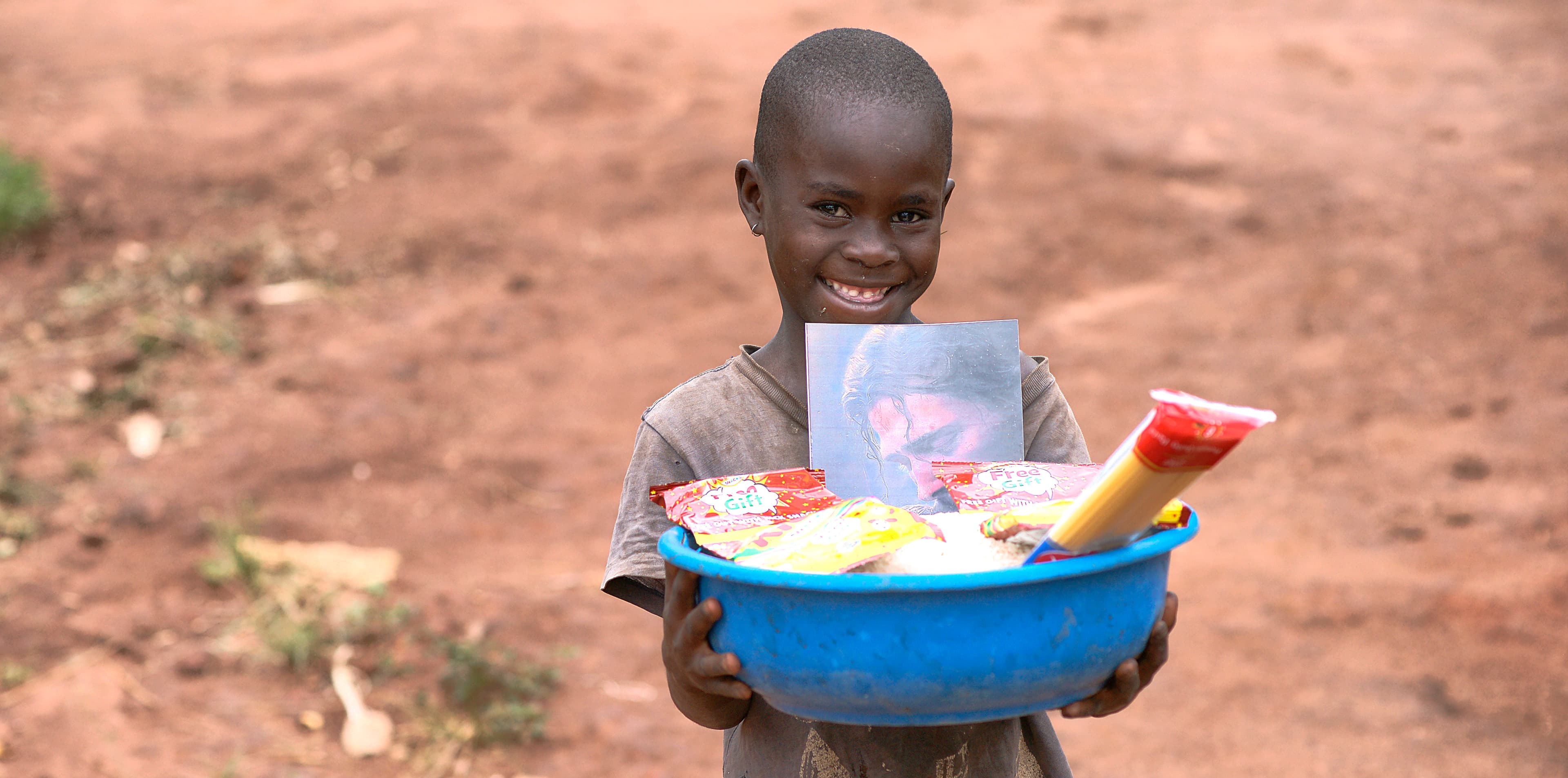 A boy stands outdoors holding a blue basin containing gospel tracts and a booklet.