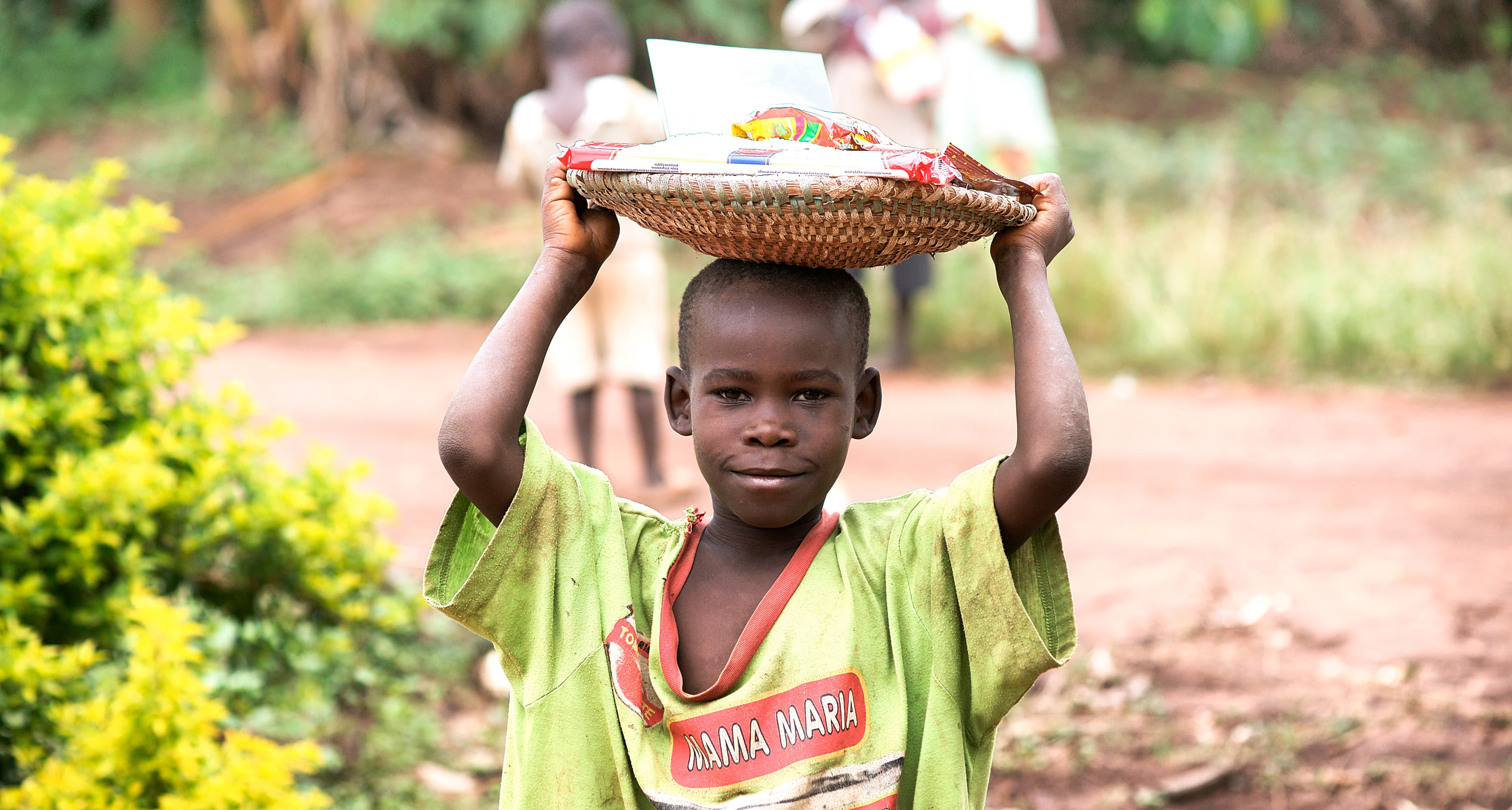 Boy carries a woven basket of food on his head near bushes.