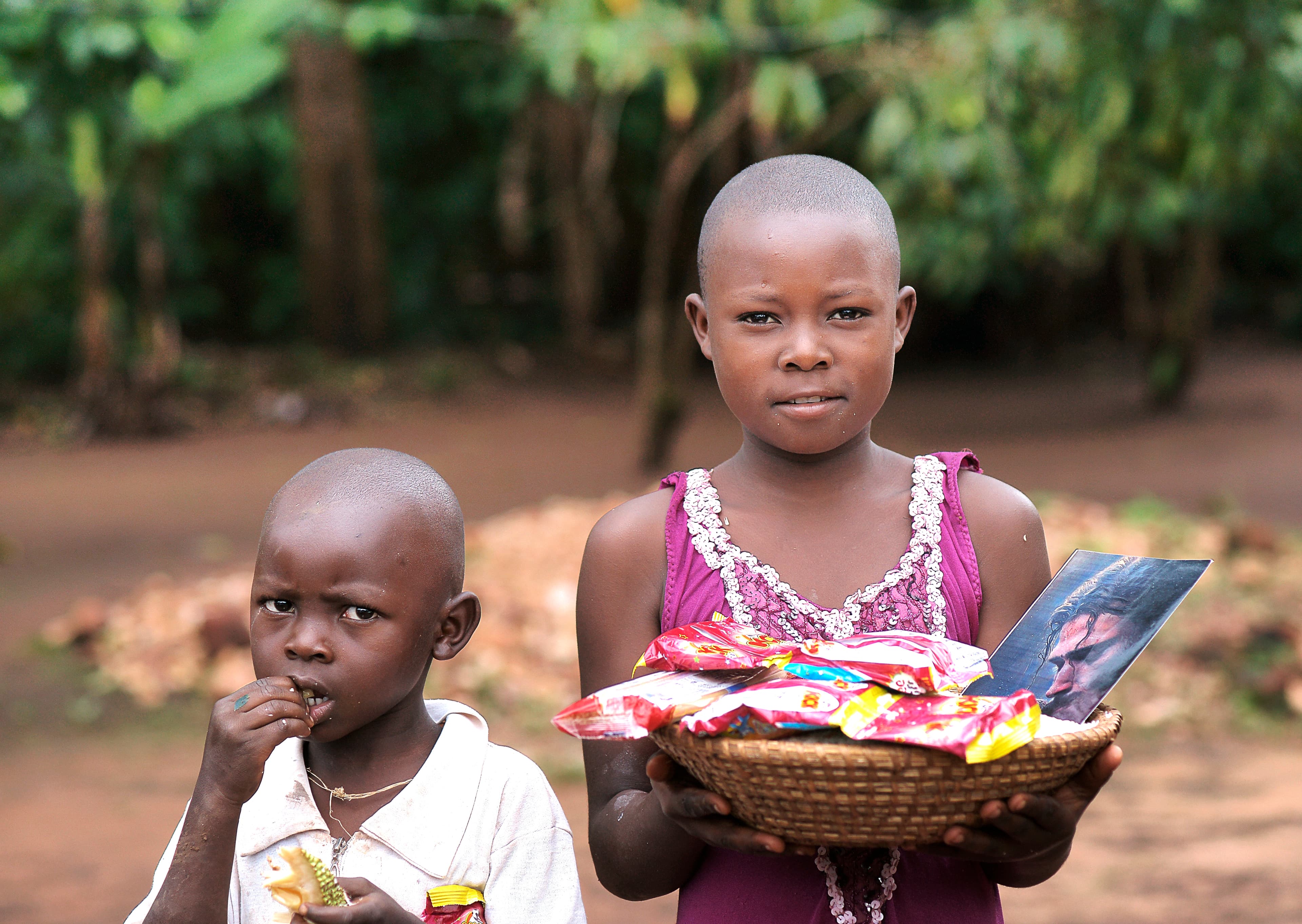 Older girl carries a woven basket with food and a photo card while a younger child stands beside her.