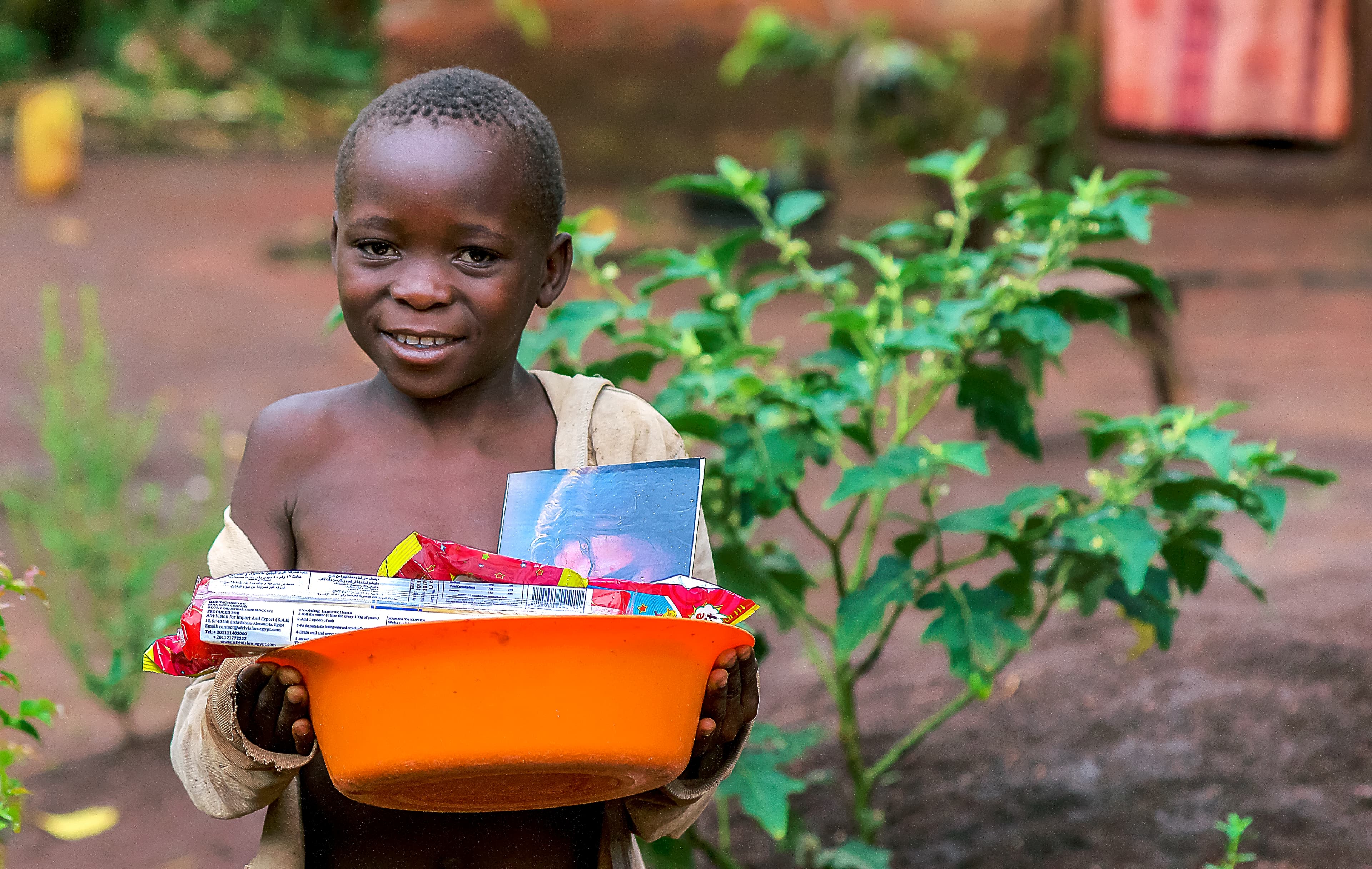 Boy stands in a field holding an orange basin filled with food items.