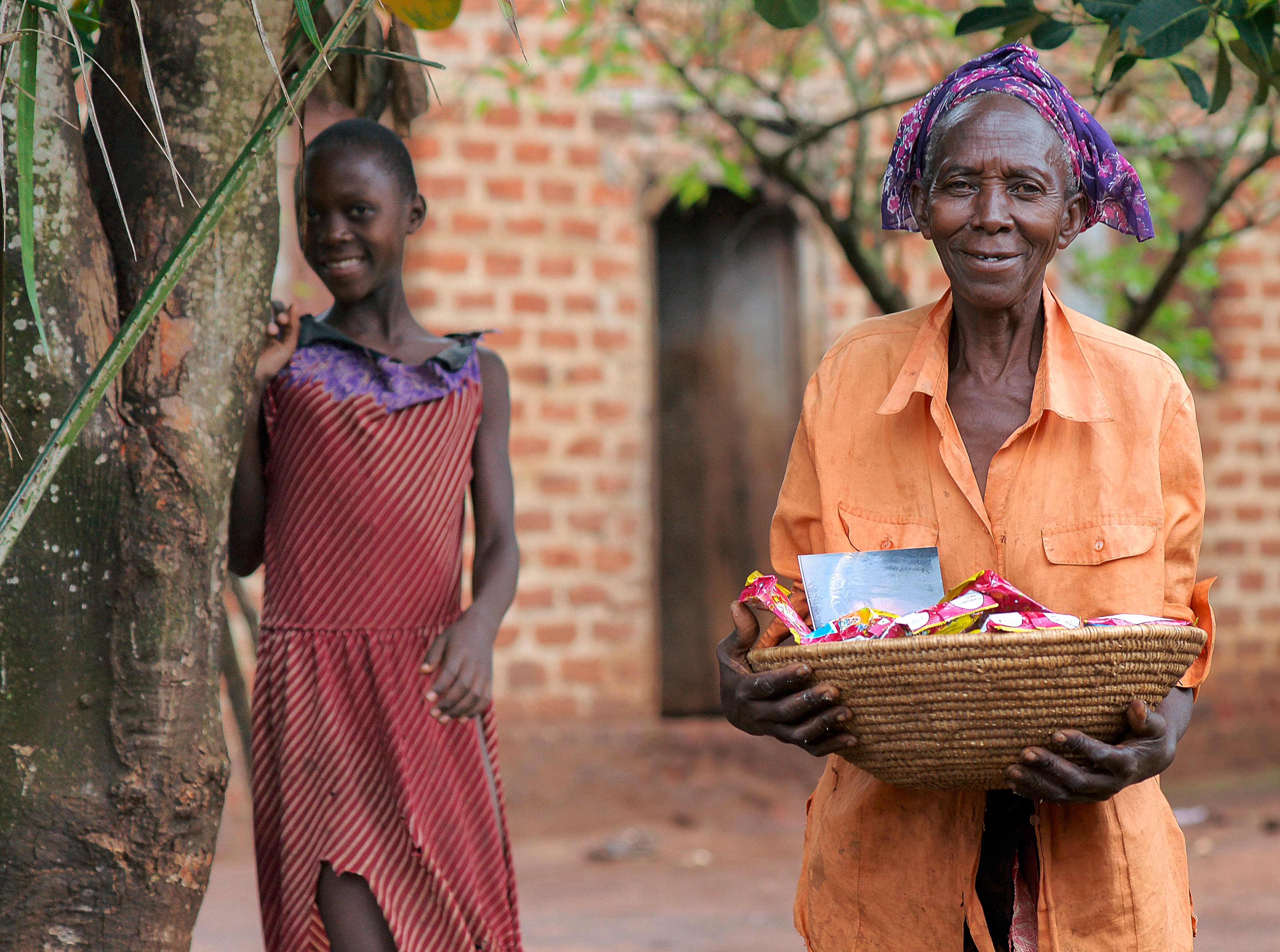 Girl and an elderly woman stand outside a mud-walled home, the girl holding a food package.