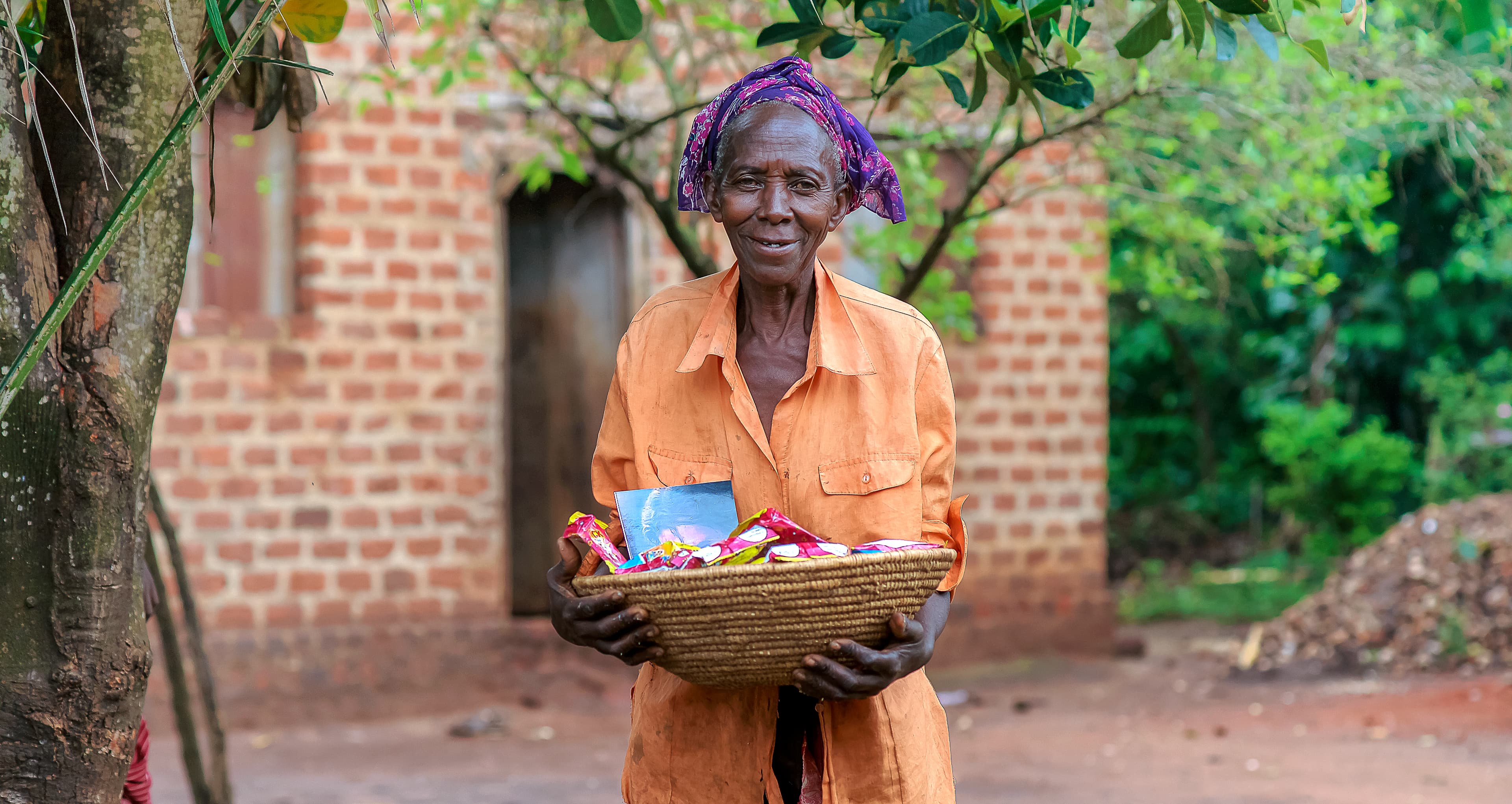 Woman in a purple headscarf stands outside a brick building holding a basket of food.