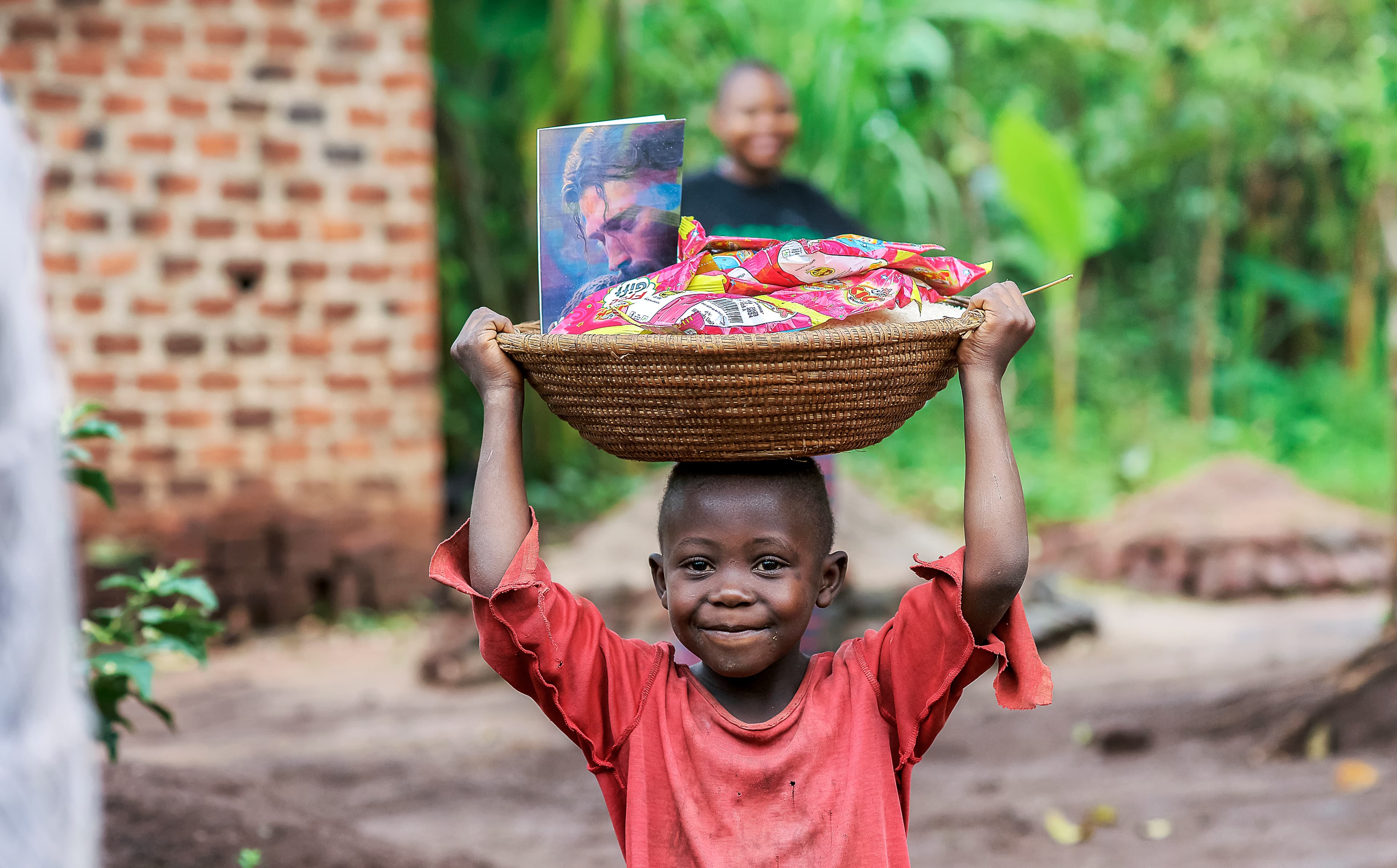 Boy carries a woven basket with food and a photo card on his head near a shelter.