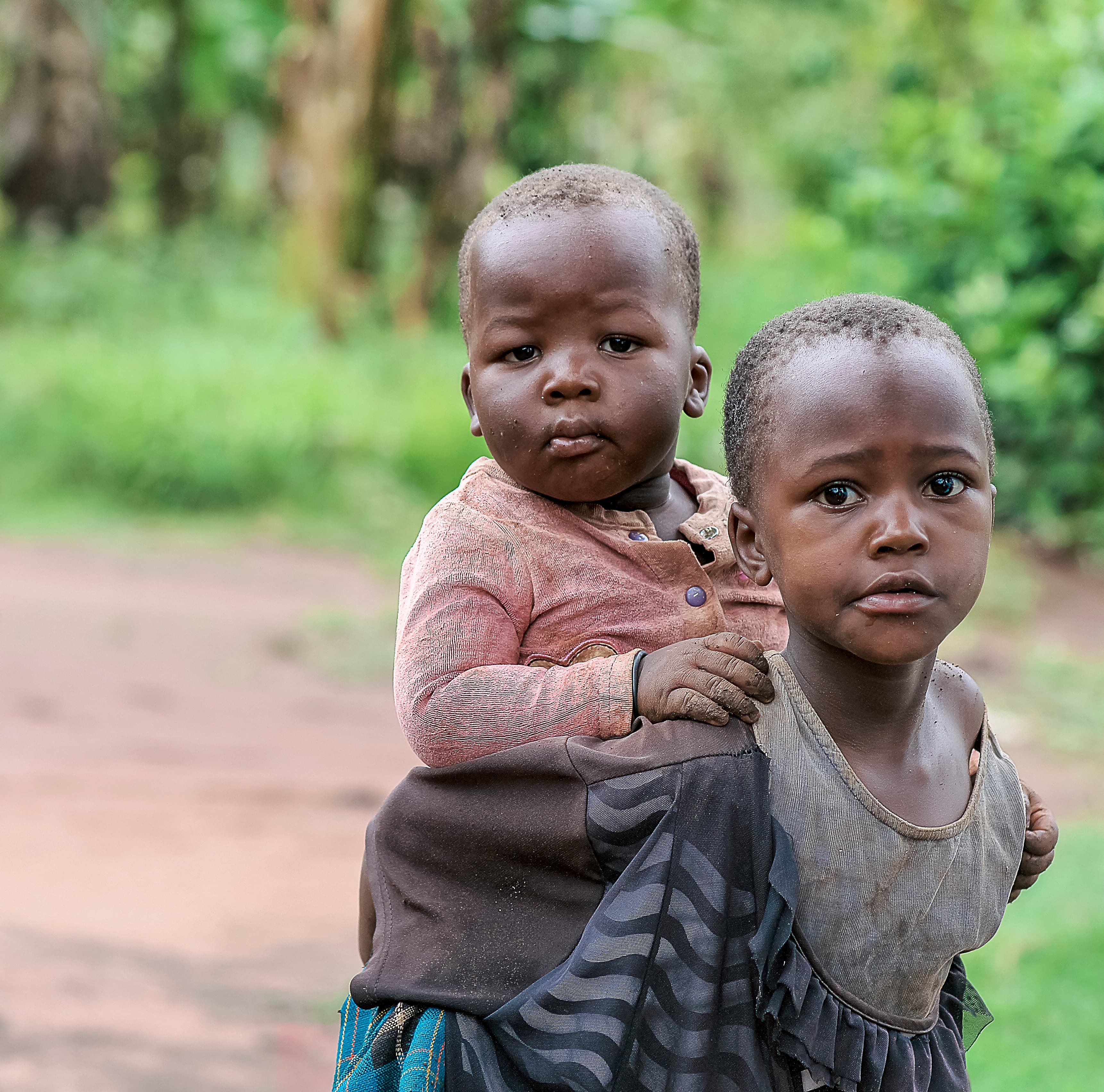 Two young children stand on a dirt path, one carrying the other on her hip.